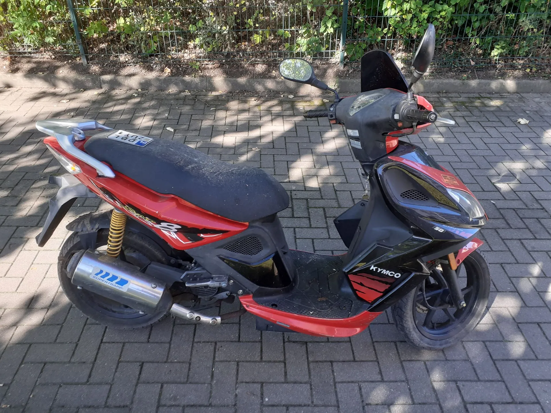 Czerwono-czarny skuter marki Kymco zaparkowany na kostce brukowej. Pojazd ma czarne siedzenie, srebrny wydech z napisem 'SUPERTRAXX' oraz widoczne logo 'KYMCO' na przedniej owiewce. W tle znajduje się zielony żywopłot i metalowy płot.