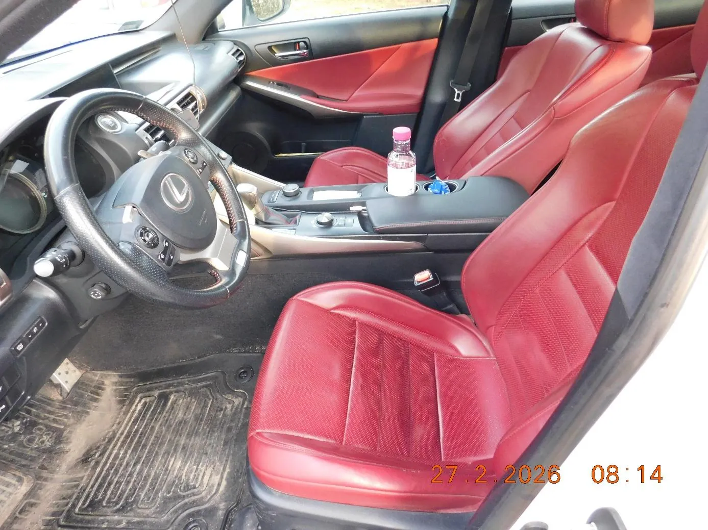 Wnętrze samochodu marki Lexus z czerwonymi skórzany siedzeniami. Widoczna kierownica z logo Lexus, czarna deska rozdzielcza, centralna konsoleta z butelką o różowej nakrętce. Na podłodze brudne dywany. W prawym dolnym rogu widoczny znaczek daty i godziny: 27.2.2026 08:14. Drzwi kierowcy są otwarte, pokazując czerwoną obicię drzwi.