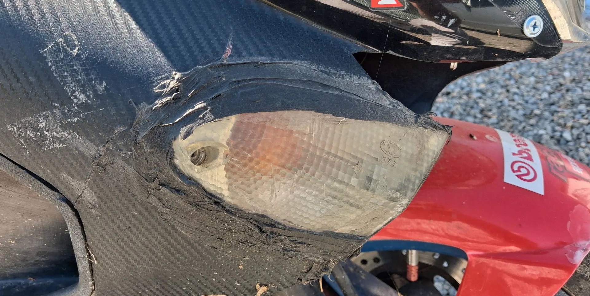 Zbliżenie na uszkodzony element karoserii motocykla. Widoczny jest czarny element z teksturą włókna węglowego, który jest pęknięty i prowizorycznie sklejony czarną taśmą wokół pomarańczowego kierunkowskazu. Obok znajduje się czerwony element plastikowy z fragmentem białego logo.