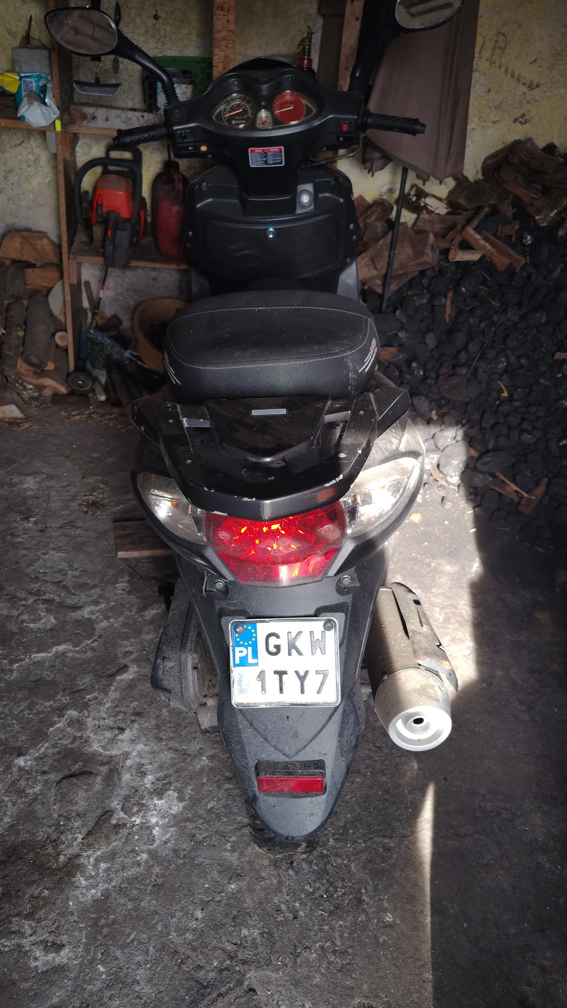 Czarny skuter widziany z góry, ustawiony poziomo. Tablica rejestracyjna z napisem 'PL GKW 1TY7' umieszczona pionowo. Widoczne czerwone światło tylnego reflektora, wydech, kierownica z lusterkami i wskaźnikami. W tle narzędzia, drewno i betonowa podłoga.