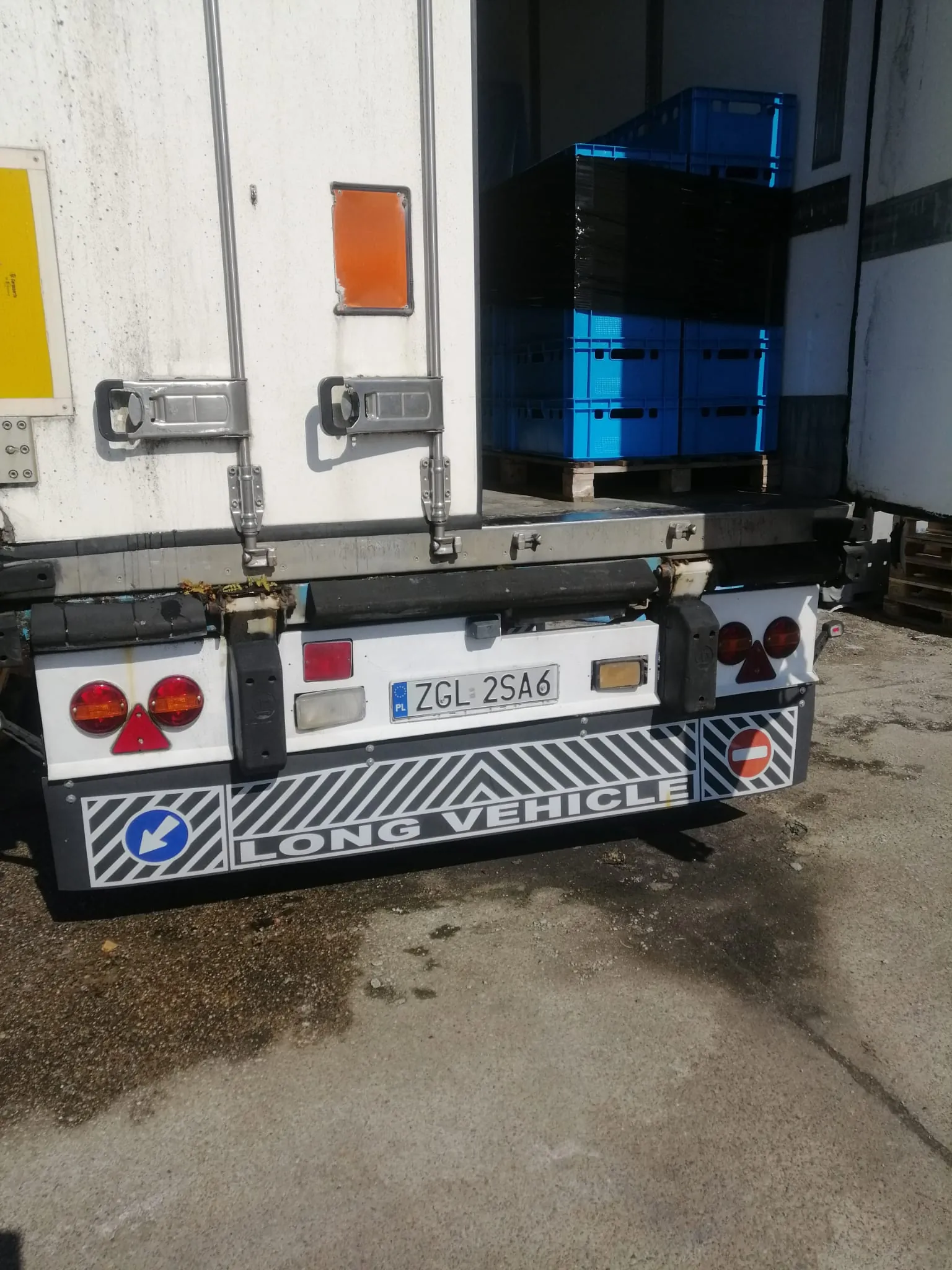 Tylna część białej naczepy ciężarówki z otwartymi drzwiami, wewnątrz których widoczne są niebieskie plastikowe skrzynie transportowe ułożone na palecie. Na zderzaku znajduje się polska tablica rejestracyjna o numerze ZGL 2SA6, światła tylne oraz biało-szary pas z napisem LONG VEHICLE i znakami ostrzegawczymi.