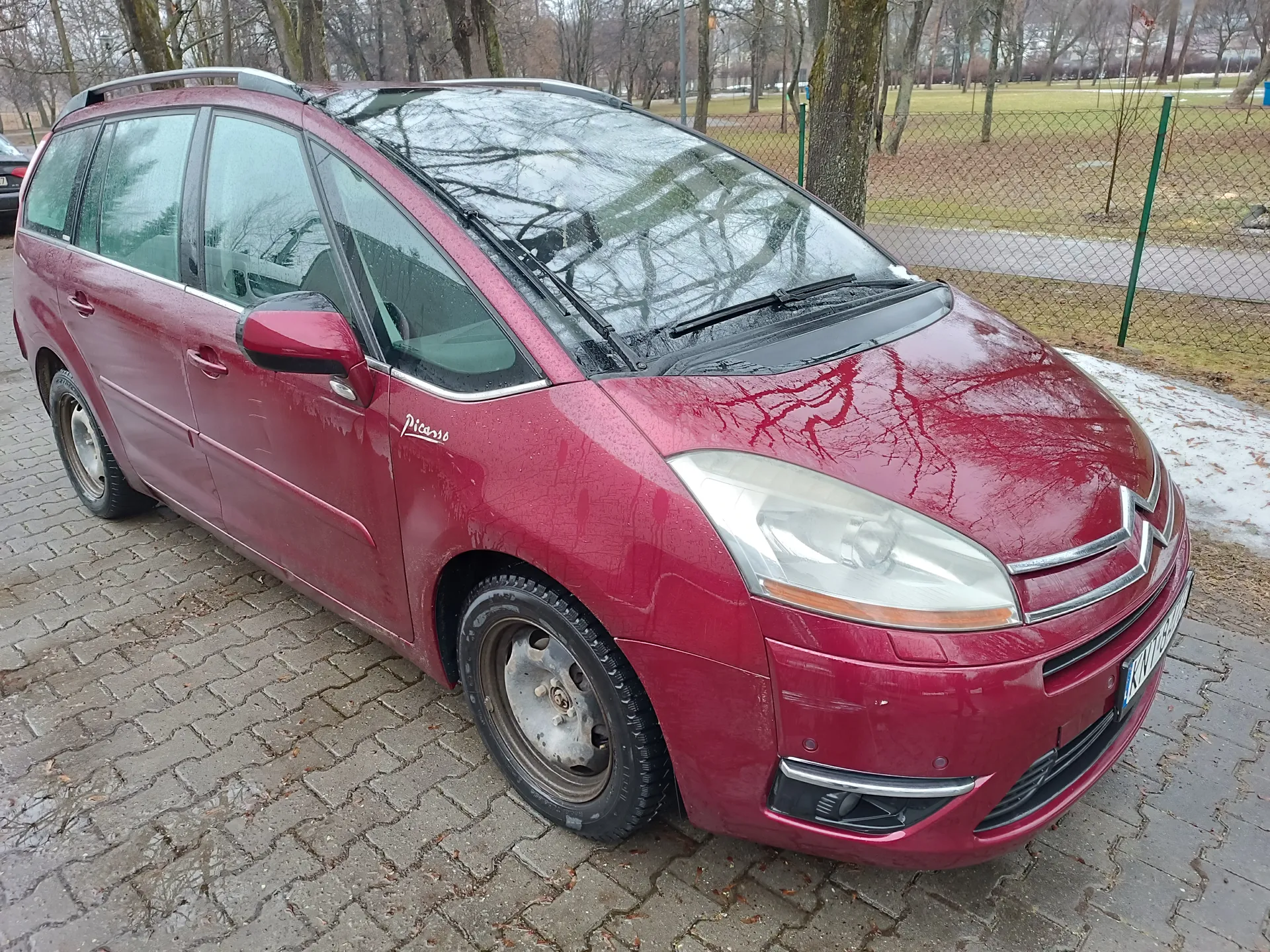 Czerwony samochód Citroën Picasso zaparkowany na brukowanej powierzchni. W tle siatka ogrodzeniowa, drzewa bez liści, trawa i resztki śniegu. Na boku pojazdu widoczny napis „Picasso”. Opony mają ślady zużycia, a na szybie odbijają się gałęzie drzew.