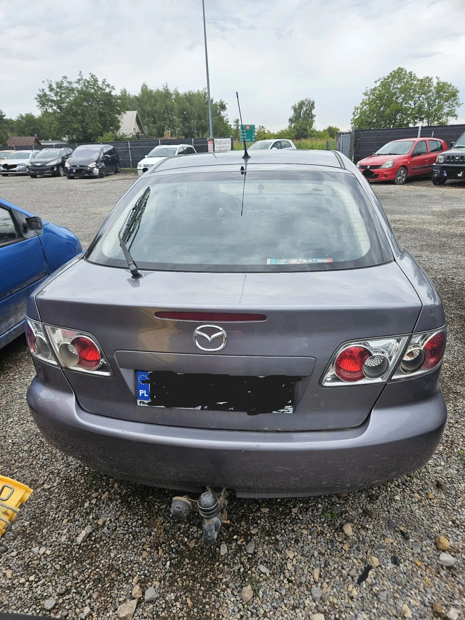 Tylny widok szarego samochodu marki Mazda zaparkowanego na żwirowym placu. Samochód posiada hak holowniczy oraz polską tablicę rejestracyjną, której numery są zakryte. W tle widoczne są inne zaparkowane samochody w różnych kolorach oraz zielone drzewa pod jasnym, zachmurzonym niebem.