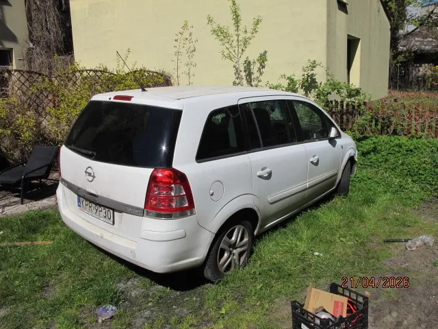 Biały samochód Opel typu kombi zaparkowany na trawie, widok od tyłu. Na tablicy rejestracyjnej widoczny numer K-ER-930. W tle żółty budynek, zielone krzewy, ogrodzenie i czarne leżaki. W prawym dolnym rogu obrazu data 21/04/2026 oraz czarna skrzynka z przedmiotami. Na trawie rozrzucone są różne przedmioty.