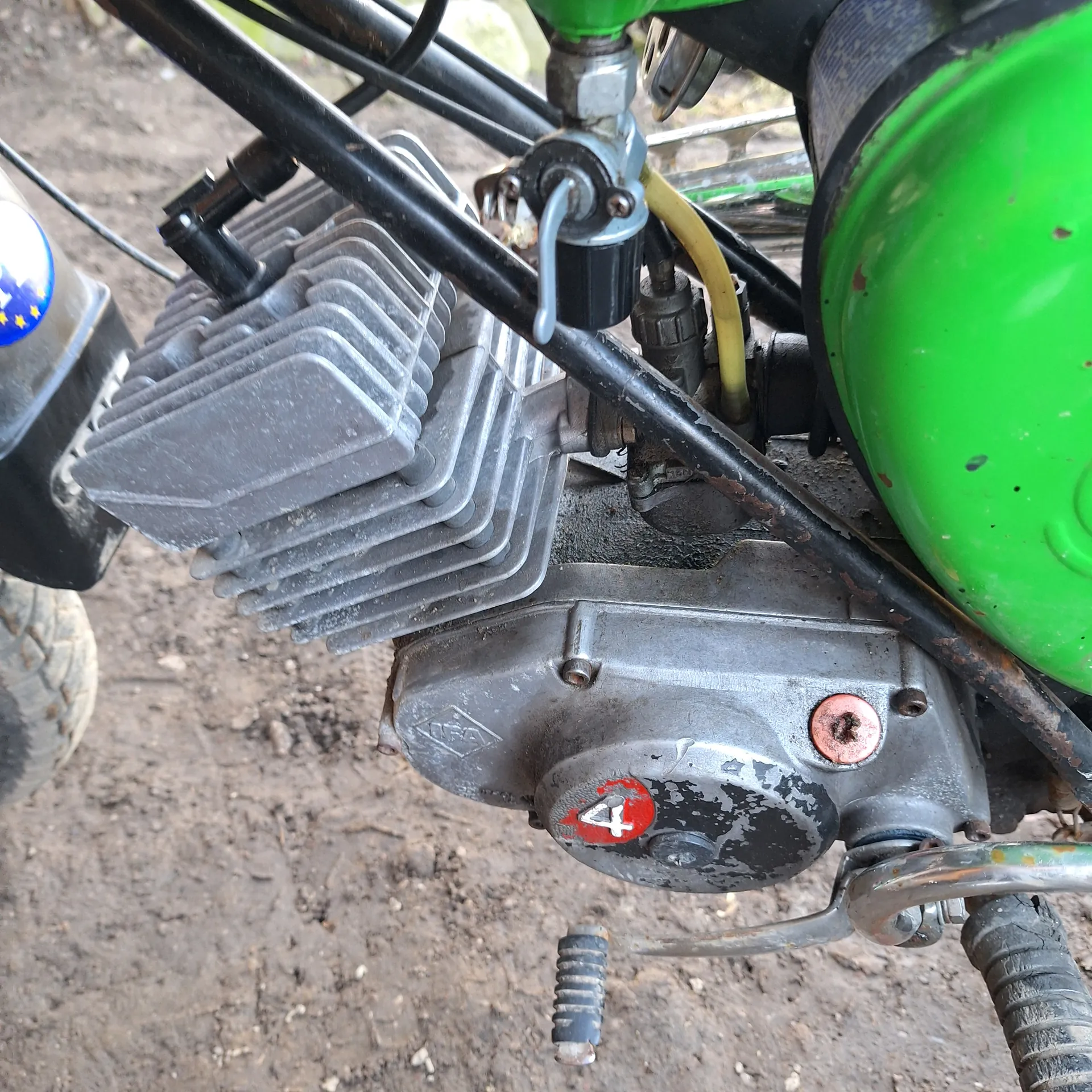 Zbliżenie na silnik motocykla z zielonym zbiornikiem paliwa w lewym górnym rogu. Widoczne są metalowe, żeberkowane elementy chłodzenia silnika, czarne przewody, żółty wąż oraz czerwony okrągły element z białym symbolem. Na tle brudna, ziemista powierzchnia. W dolnej części obrazu fragment opony z niebieską naklejką z żółtymi gwiazdkami.