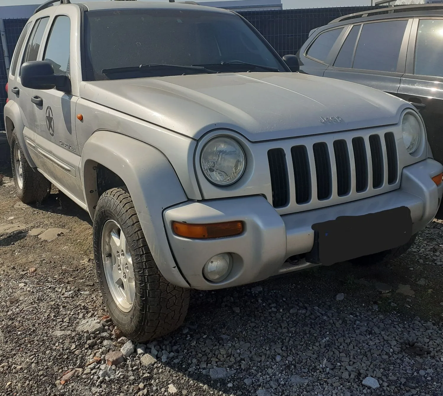 Srebrny samochód terenowy marki Jeep zaparkowany na żwirowym podłożu. Widoczny jest przód pojazdu z charakterystycznym grillem z siedmioma pionowymi szczelinami, okrągłymi reflektorami oraz pomarańczowymi kierunkowskazami w zderzaku. Na drzwiach kierowcy znajduje się ciemna naklejka w kształcie gwiazdy oraz napis 'LIBERTY'. W tle widoczny jest fragment innego, ciemnego samochodu.