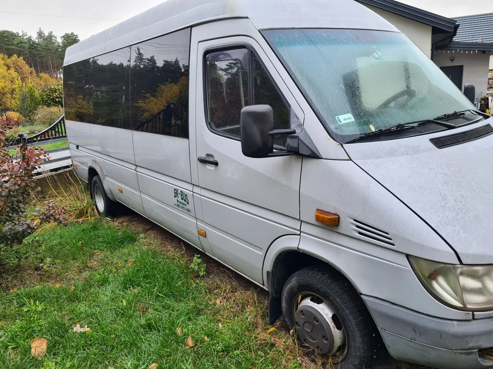 Srebrny autobus typu van zaparkowany na trawie. Na boku pojazdu widoczny zielony napis "SK-BUS" oraz mniejszy tekst poniżej. W tle dom z ciemnym dachem, drzewa z jesienną roślinnością oraz zarośla. Przednia szyba i lusterko boczne są widoczne, a na trawie leżą opadłe liście.