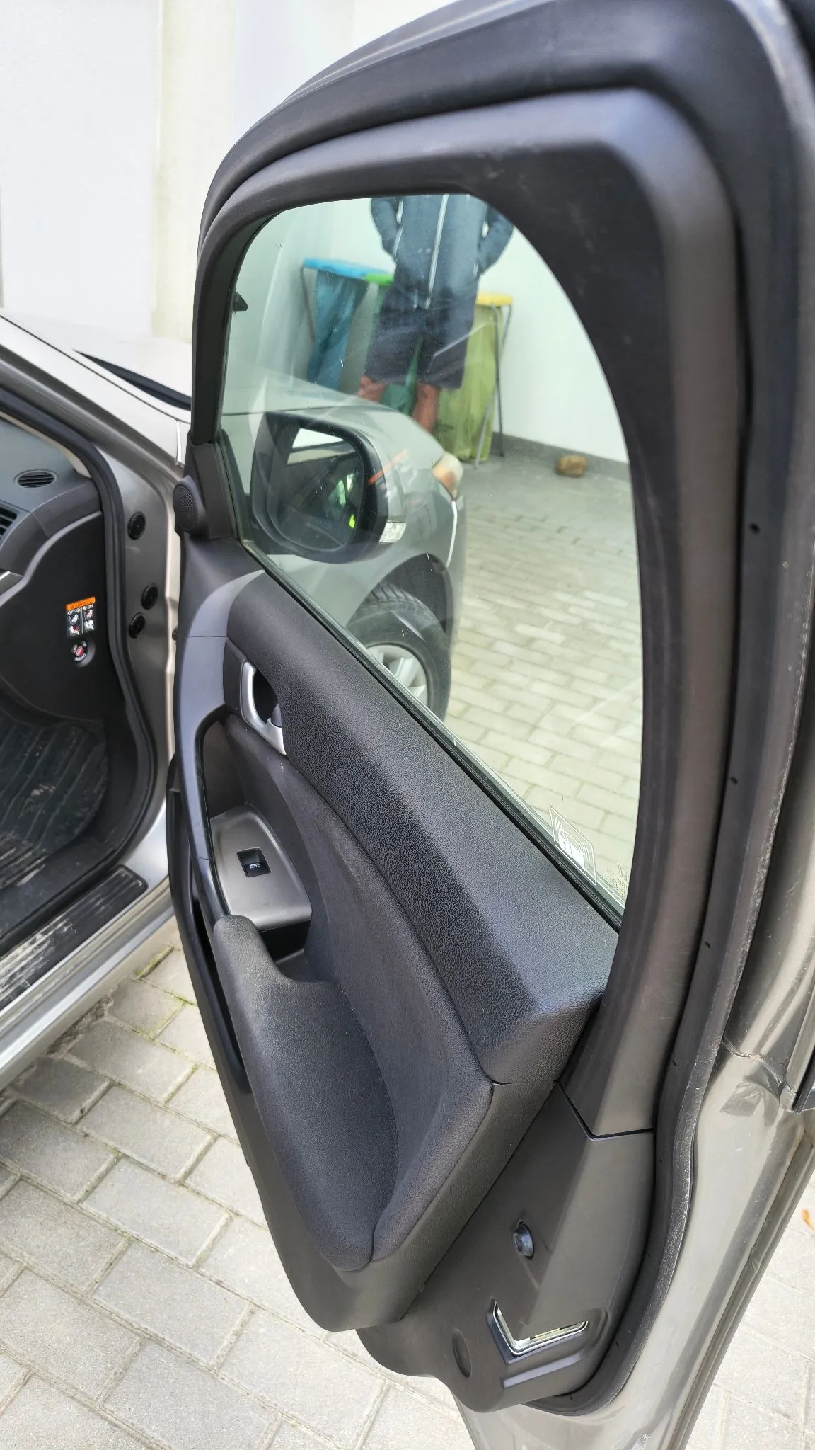Zbliżenie na otwarte drzwi samochodu od strony wnętrza. Widoczny jest czarny plastikowy panel drzwi z uchwytem i szybą, w której odbija się postać osoby oraz fragment podjazdu z kostki brukowej.