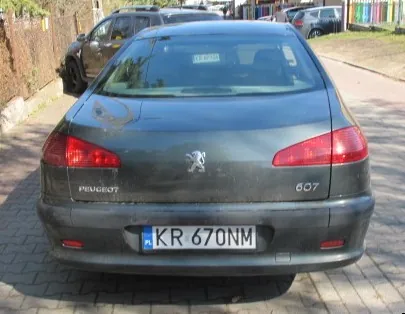 Tył ciemnoszarego samochodu Peugeot 607 zaparkowanego na brukowanej ulicy. Widoczne są czerwone światła tylnego, logo marki, napis 'PEUGEOT' po lewej stronie, numer modelu '607' po prawej stronie oraz tablica rejestracyjna 'KR 670NM' z niebieskim pasem po lewej stronie. W tle znajdują się inne zaparkowane pojazdy, zieleń i budynki.
