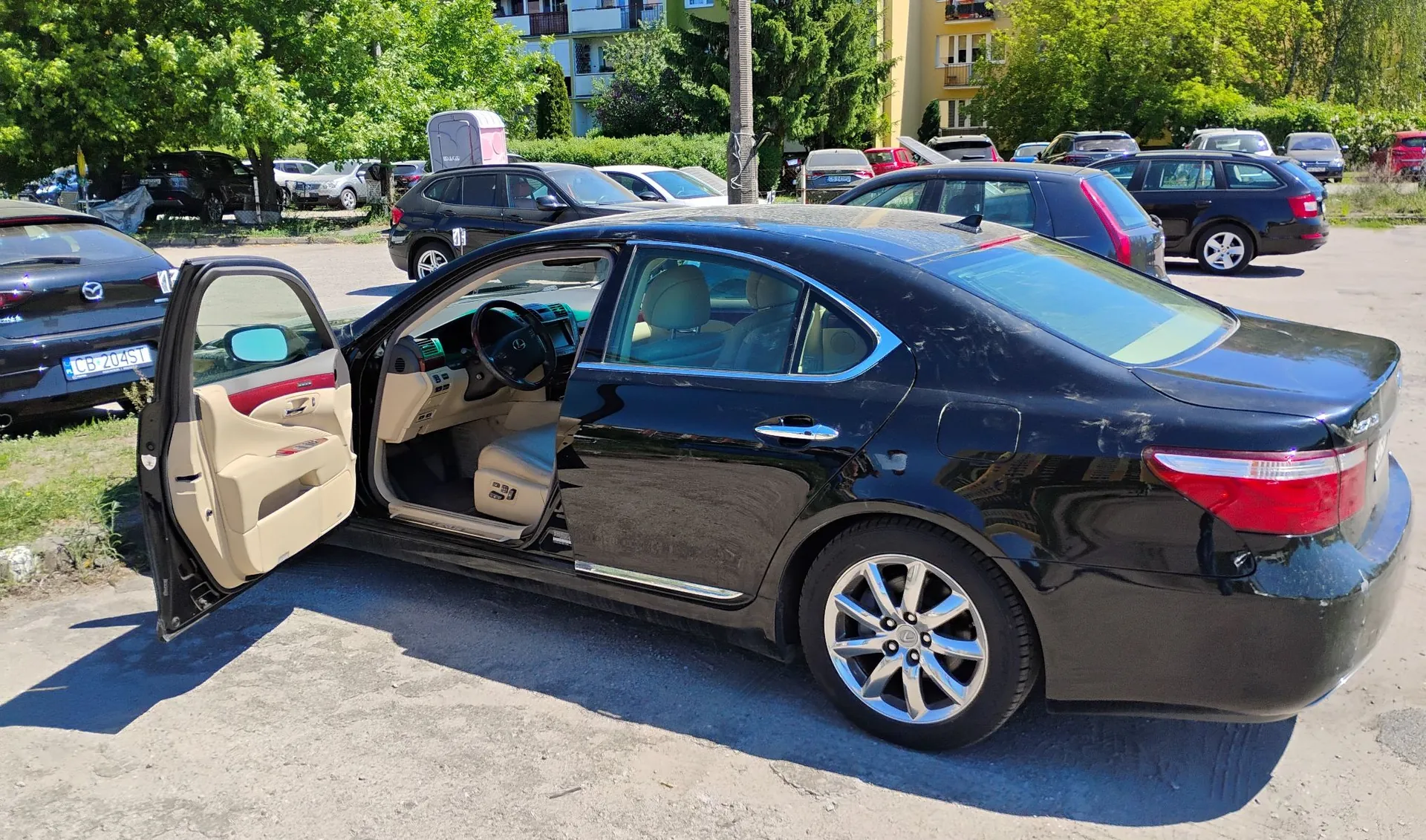 Czarny samochód osobowy typu sedan zaparkowany na utwardzonym placu. Przednie lewe drzwi kierowcy są otwarte, odsłaniając beżową tapicerkę wnętrza. Na tylnym błotniku i klapie bagażnika widoczne są zarysowania i uszkodzenia lakieru. W tle widać inne zaparkowane samochody, zielone drzewa oraz fragmenty budynków mieszkalnych.