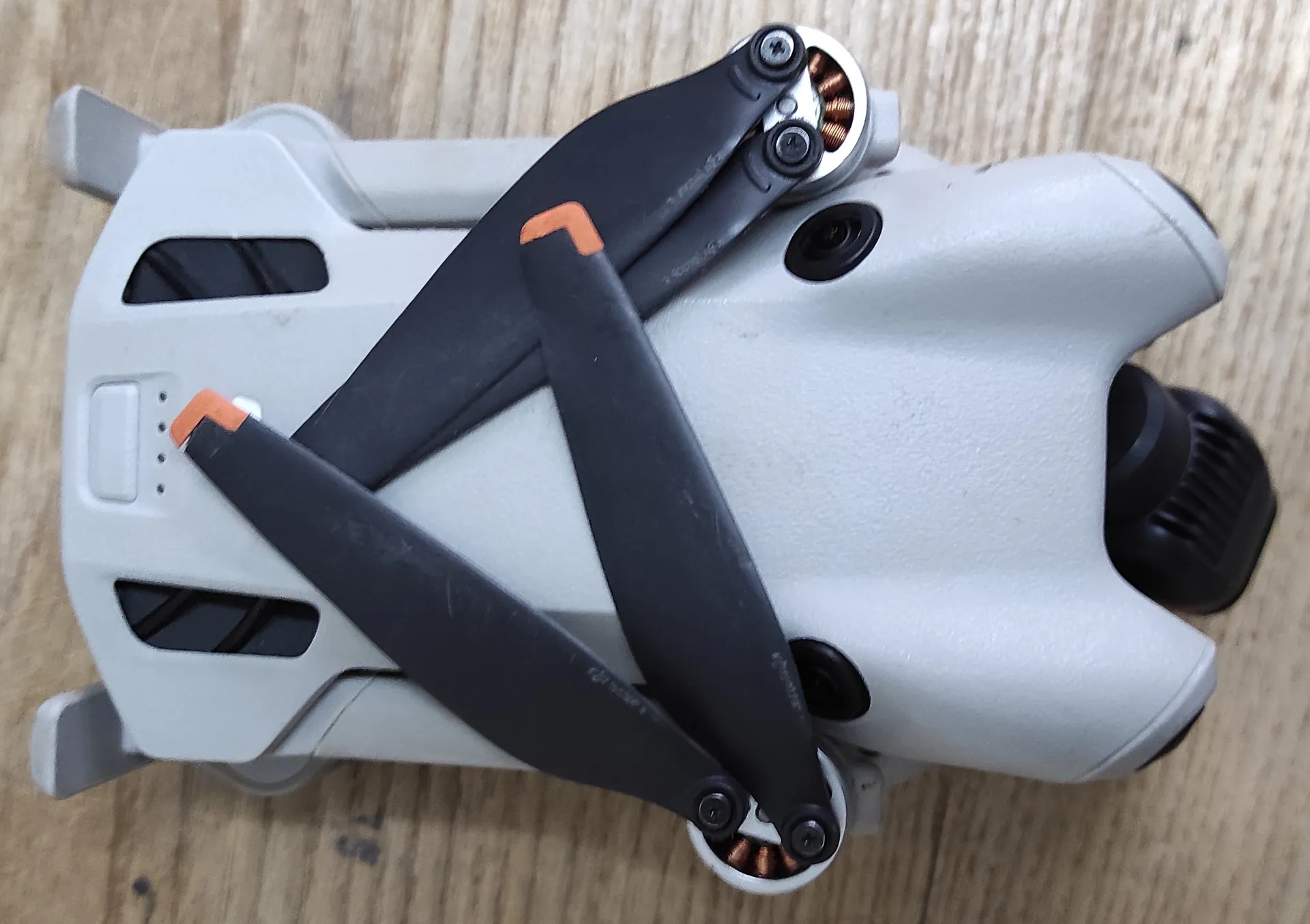 Widok z góry na biały dron z czterema czarnymi śmigłami, leżący na drewnianej powierzchni. Dron ma obły kształt z widocznymi silnikami na końcach ramion oraz czarnym elementem z przodu.