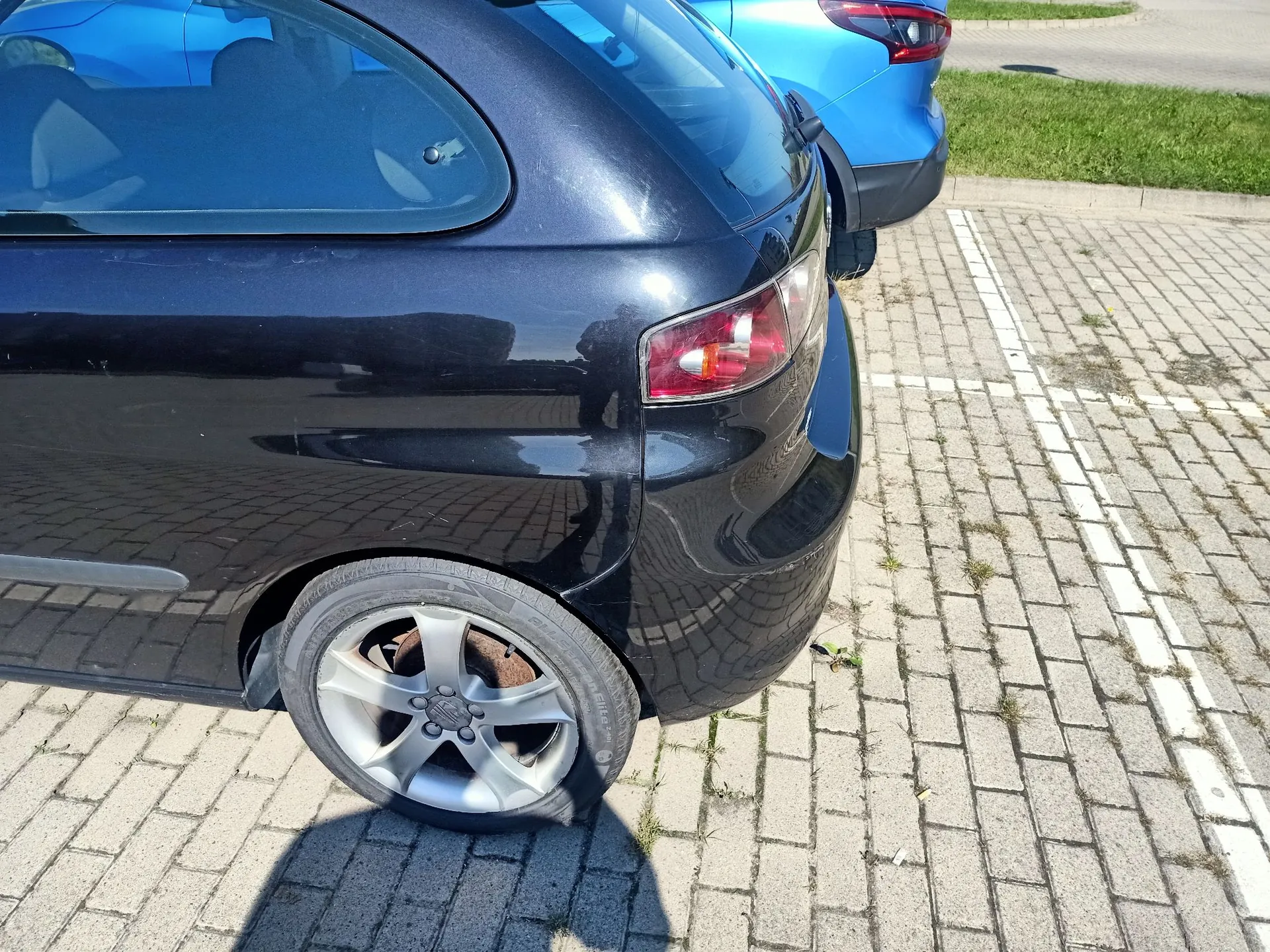Zbliżenie na tył czarnego samochodu zaparkowanego na kostce brukowej. Widoczny jest tylny lewy błotnik, lampa tylna oraz koło z pięcioramienną felgą aluminiową. W tle widoczny jest fragment innego, niebieskiego samochodu oraz trawnik.