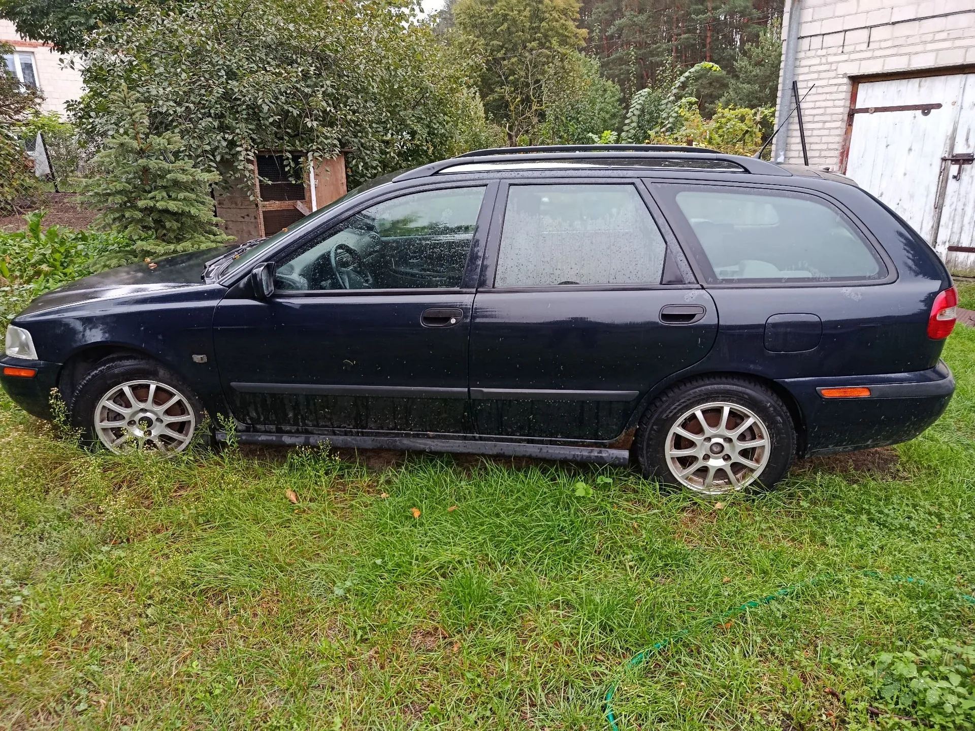 Czarny samochód typu kombi zaparkowany na trawie przed budynkiem i drzewami. Pojazd jest brudny, widoczne są na nim zacieki i pył. Samochód ma srebrne felgi aluminiowe z wieloma szprychami.
