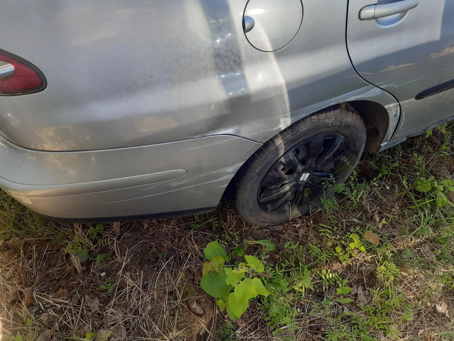 Zbliżenie na tylną, prawą część srebrnego samochodu. Widoczny jest tylny zderzak, błotnik oraz koło z czarną felgą. Samochód stoi na nieutwardzonym terenie z trawą i suchymi liśćmi.