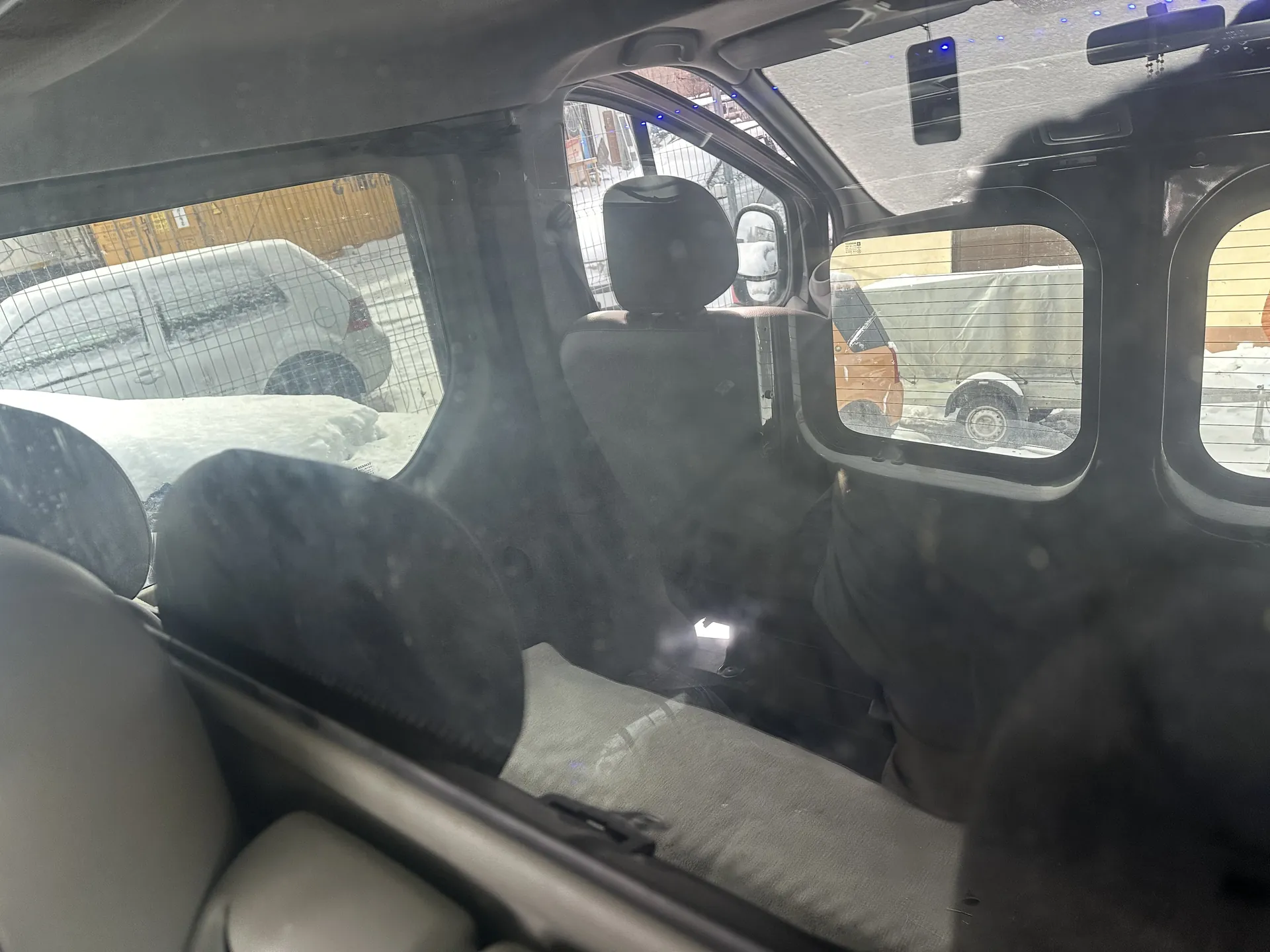 Wnętrze samochodu widziane z tyłu, z widocznymi siedzeniami, zagłówkami i siatką oddzielającą przestrzeń. Przez okna widać zewnętrzne elementy: biały samochód, pomarańczowy pojazd, budynki oraz śnieg na ziemi. Na zewnątrz panuje zimowa pogoda, na powierzchniach widoczna warstwa śniegu. W samochodzie widoczne są również lusterko wsteczne i elementy wyposażenia wnętrza.