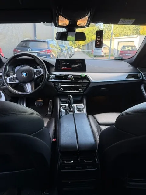 Wnętrze samochodu marki BMW widziane z perspektywy tylnego siedzenia. Widoczna jest czarna skórzana tapicerka, kierownica z logo BMW, deska rozdzielcza z ekranem multimedialnym oraz konsola środkowa z lewarkiem zmiany biegów. Przez przednią szybę widoczny jest parking z zaparkowanymi samochodami, w tym niebieski samochód marki Mazda.