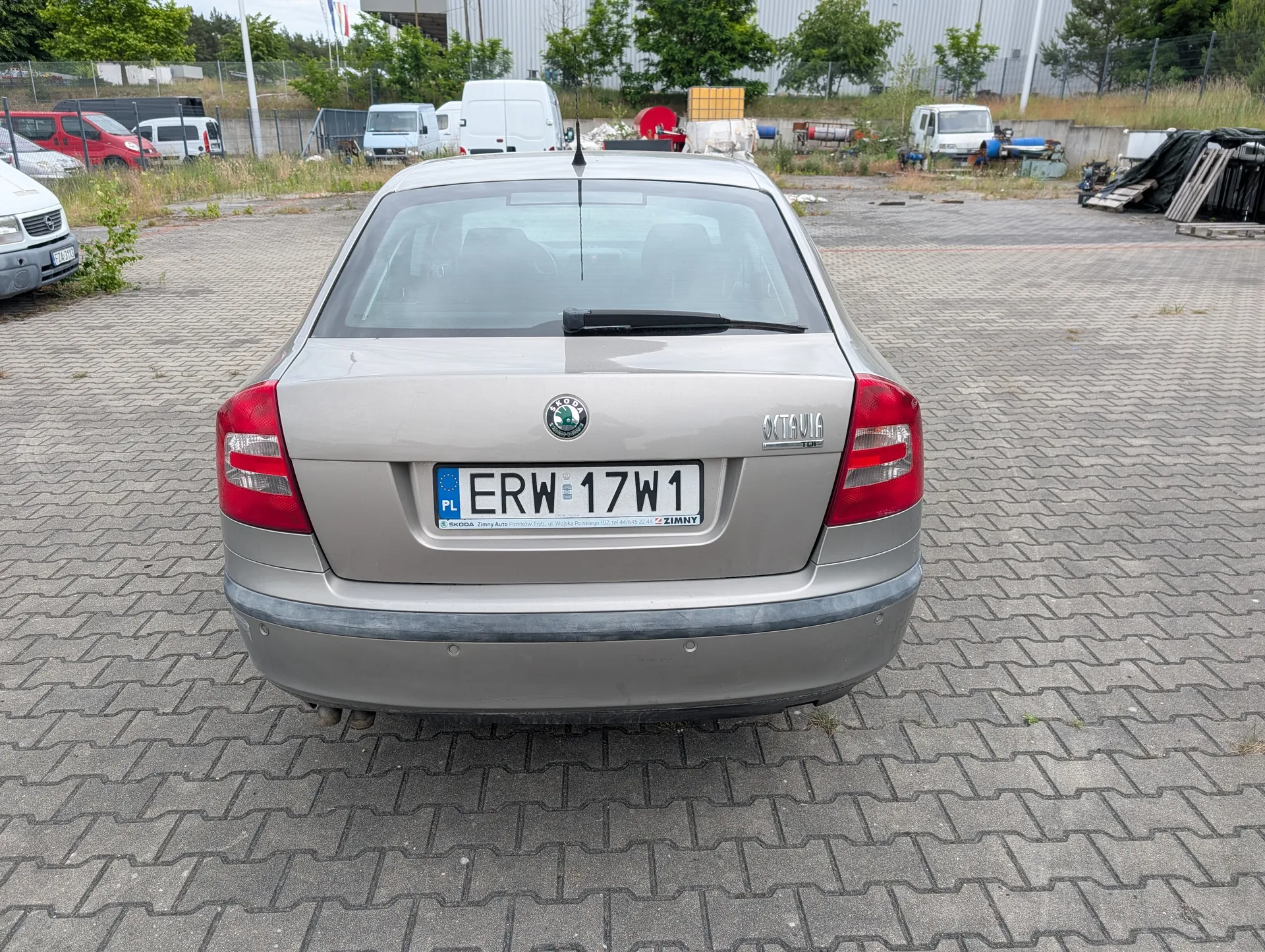 Z tyłu srebrny samochód marki Škoda Octavia z tablicą rejestracyjną PL ERW 17W1. Na tle widoczne są inne pojazdy (czerwony samochód, białe furgony), przemysłowe budynki, drzewa i ogrodzenie. Samochód stoi na wybrukowanym placu. Na tablicy rejestracyjnej widoczny napis "ZIMNY" oraz logo Škoda na bagażniku.