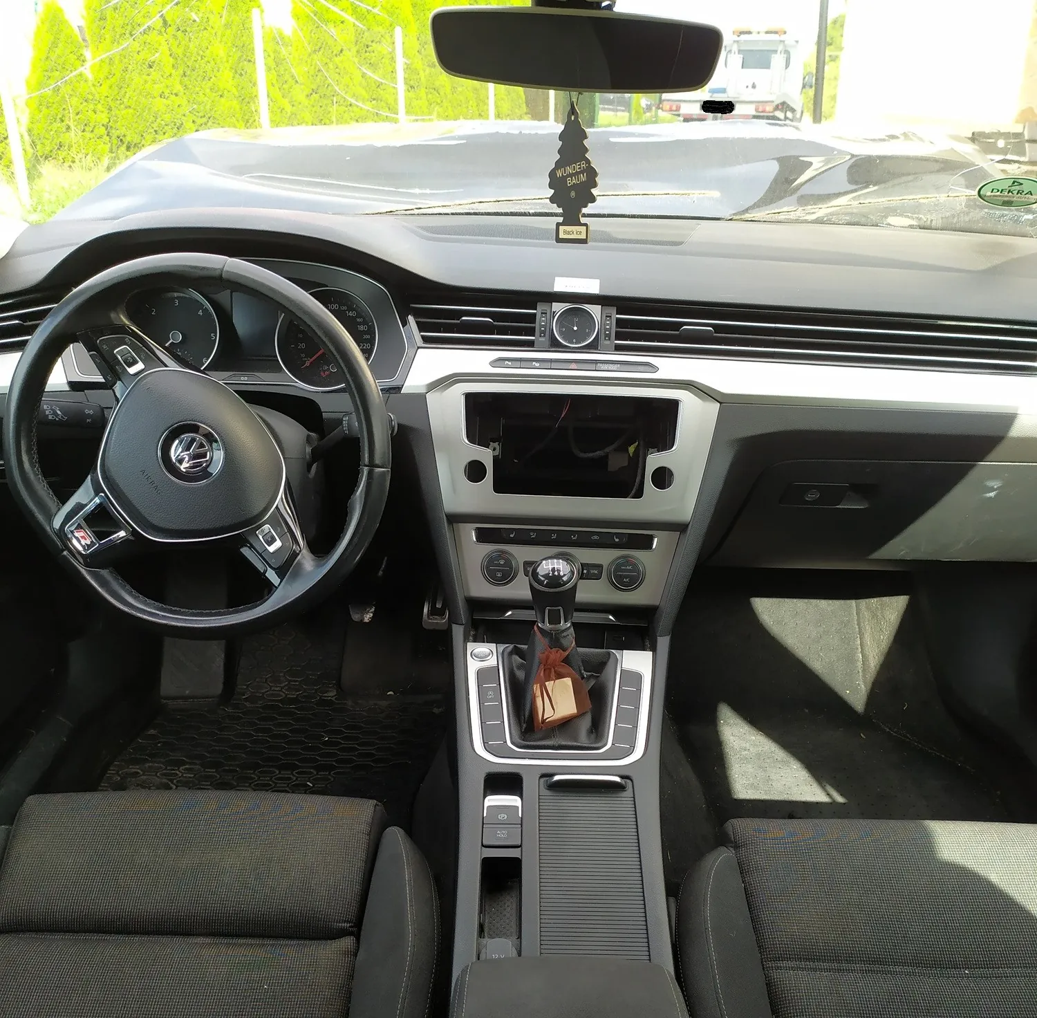 Wnętrze samochodu marki Volkswagen widziane z perspektywy kierowcy. Widoczna jest czarna kierownica z logo marki, deska rozdzielcza z brakującym radiem/ekranem w konsoli centralnej, lewarek zmiany biegów oraz dwa przednie fotele z ciemnej tkaniny. Na lusterku wstecznym wisi zapach samochodowy w kształcie choinki z napisem 'WUNDER BAUM'.