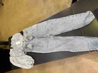 Para jasnoniebieskich dżinsów rozłożonych poziomo na ciemnej powierzchni. Dżinsy mają klasyczny kroj, widoczny jest pas z pętlami oraz biała etykieta na lewej stronie pasa. Tło składa się z żółtawej podłogi i części ciemnego mebla.