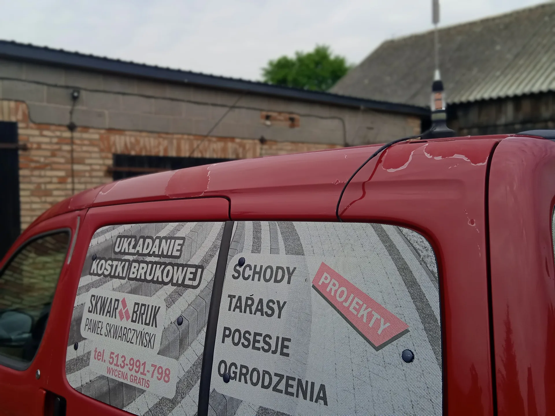 Czerwony samochód dostawczy z transparentem na tylnej szybie. Na transparentzie widoczny tekst: 'UKŁADANIE KOSTKI BRUKOWEJ', 'SKWAR BRUK', 'PAWEŁ SKWARCZYŃSKI', 'tel. 513-991-798', 'WYCENA GRATIS', 'SCHODY', 'TARASY', 'POSESJE', 'OGRODZENIA' oraz czerwony napis 'PROJEKTY'. W tle budynki z cegły i betonu, dach z blachy oraz drzewo. Niebo jest zachmurzone.