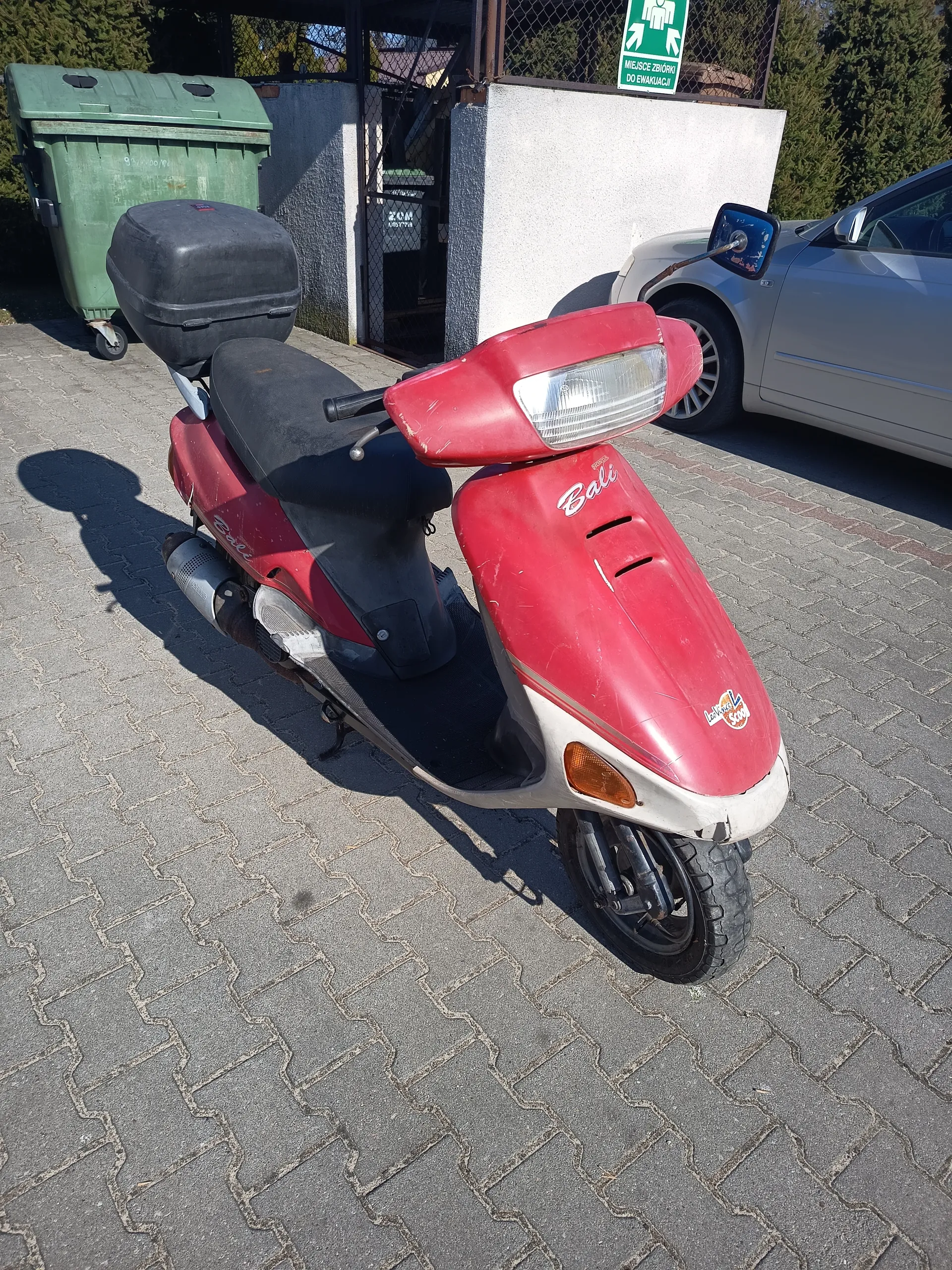 Czerwony skuter marki Bali z czarnym siedzeniem i czarnym bagażnikiem na tyle, zaparkowany na brukowanej powierzchni. W tle zielony pojemnik na śmieci, srebrny samochód, biała ściana z metalowym ogrodzeniem oraz zielony znak ewakuacyjny z napisem 'MIEJSCE ZBIÓRKI DO EWAKUACJI' i strzałkami. Skuter posiada białe napisy 'Bali' na czerwonej części oraz naklejkę z logo na przodzie.