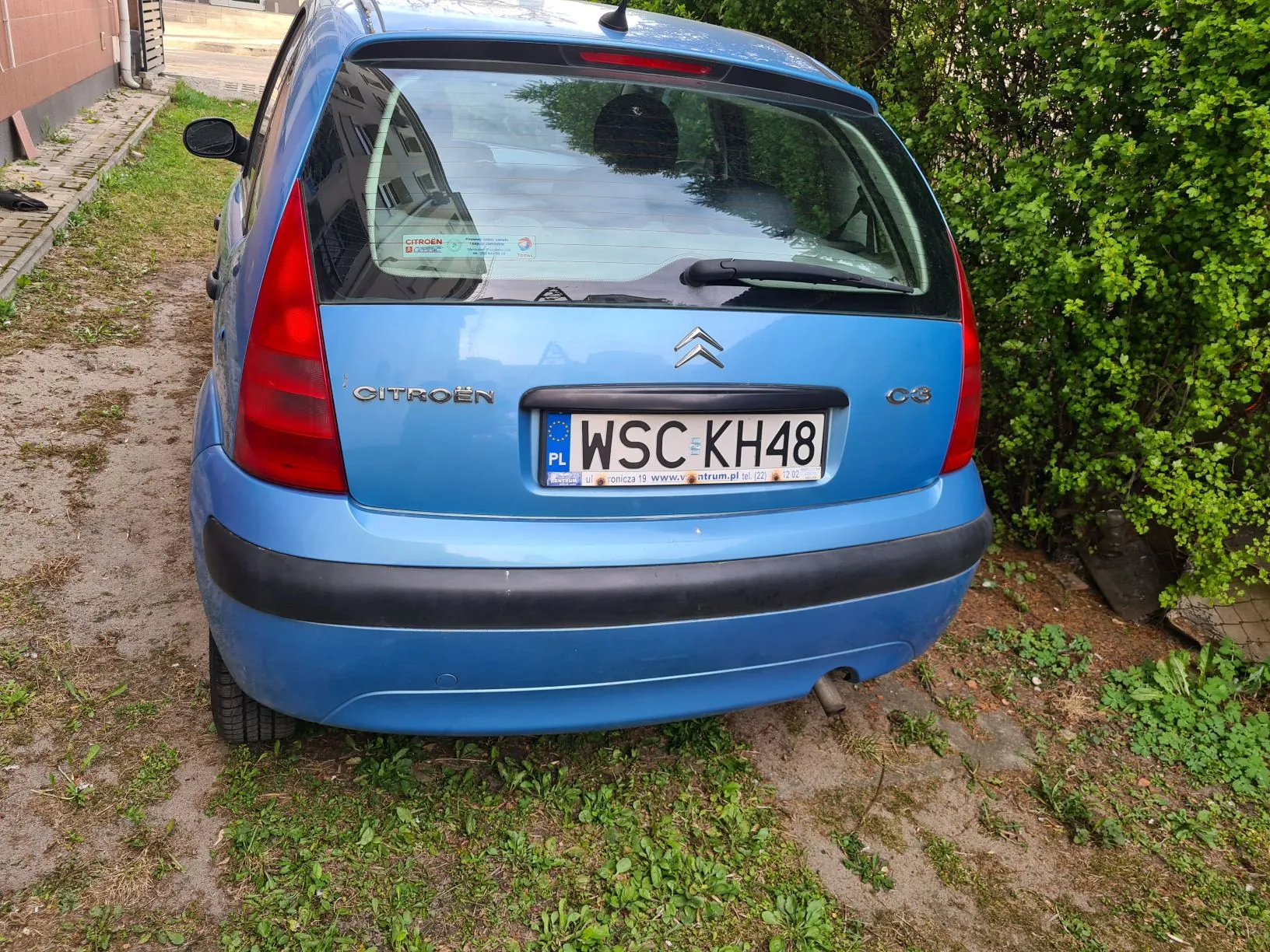 Tył niebieskiego samochodu marki Citroën C3 zaparkowanego na terenie z trawą i odsłoniętą ziemią. Widoczna tablica rejestracyjna WSC KH48 z oznaczeniem PL oraz logo marki na bagażniku. Po prawej stronie zielony krzew, po lewej budynek z ceglaną ścianą i chodnikiem. Na szybie tylnej naklejki, w tym z napisem CITROËN.