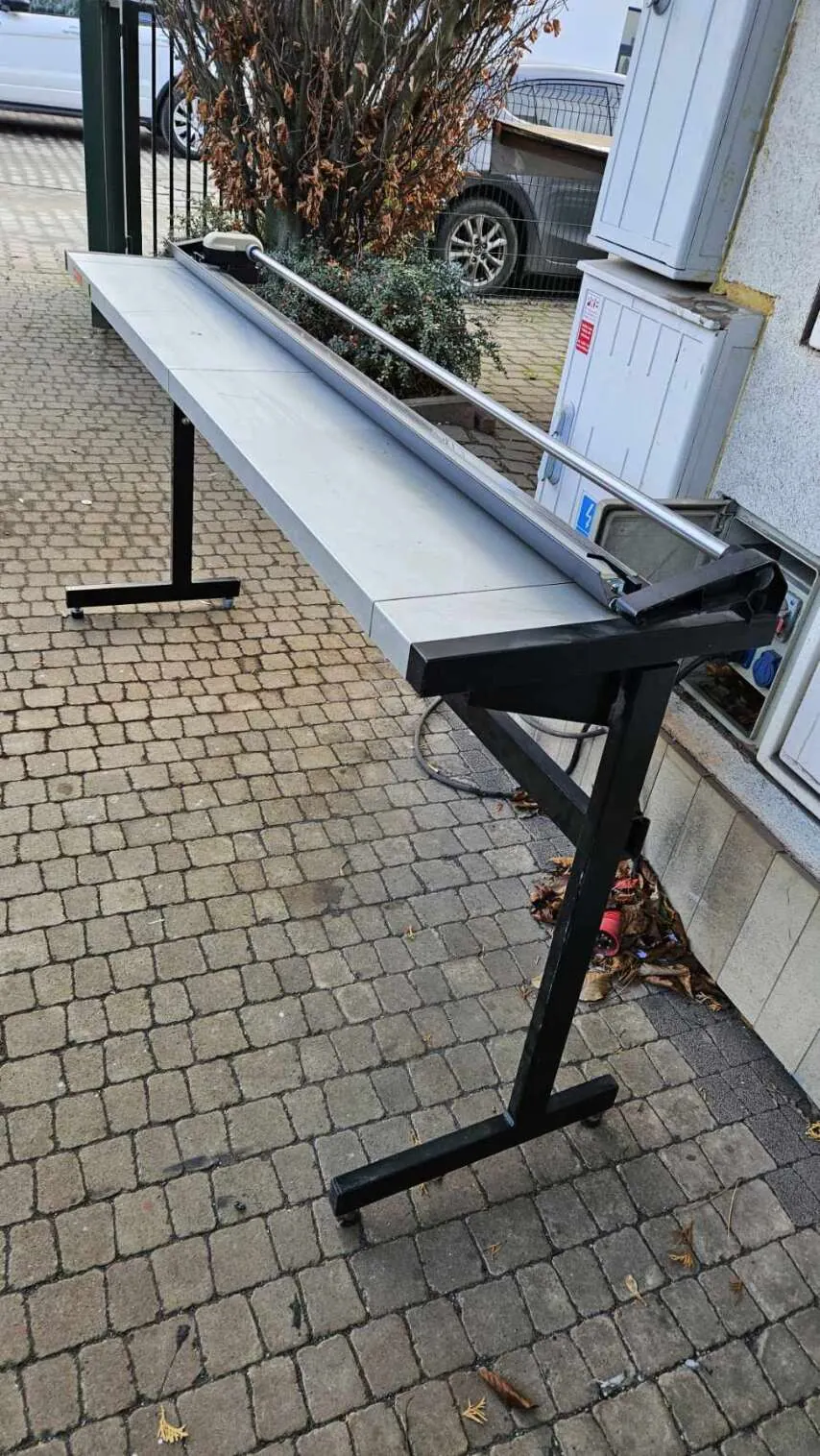 Stół z metalową ramą na kółkach umieszczony na brukowanej nawierzchni. Na stole znajduje się długie metalowe rurek. W tle widoczne są samochody, krzew z suchymi liśćmi, białe skrzynki energetyczne przy ścianie budynku oraz rozrzucone liście na ziemi.