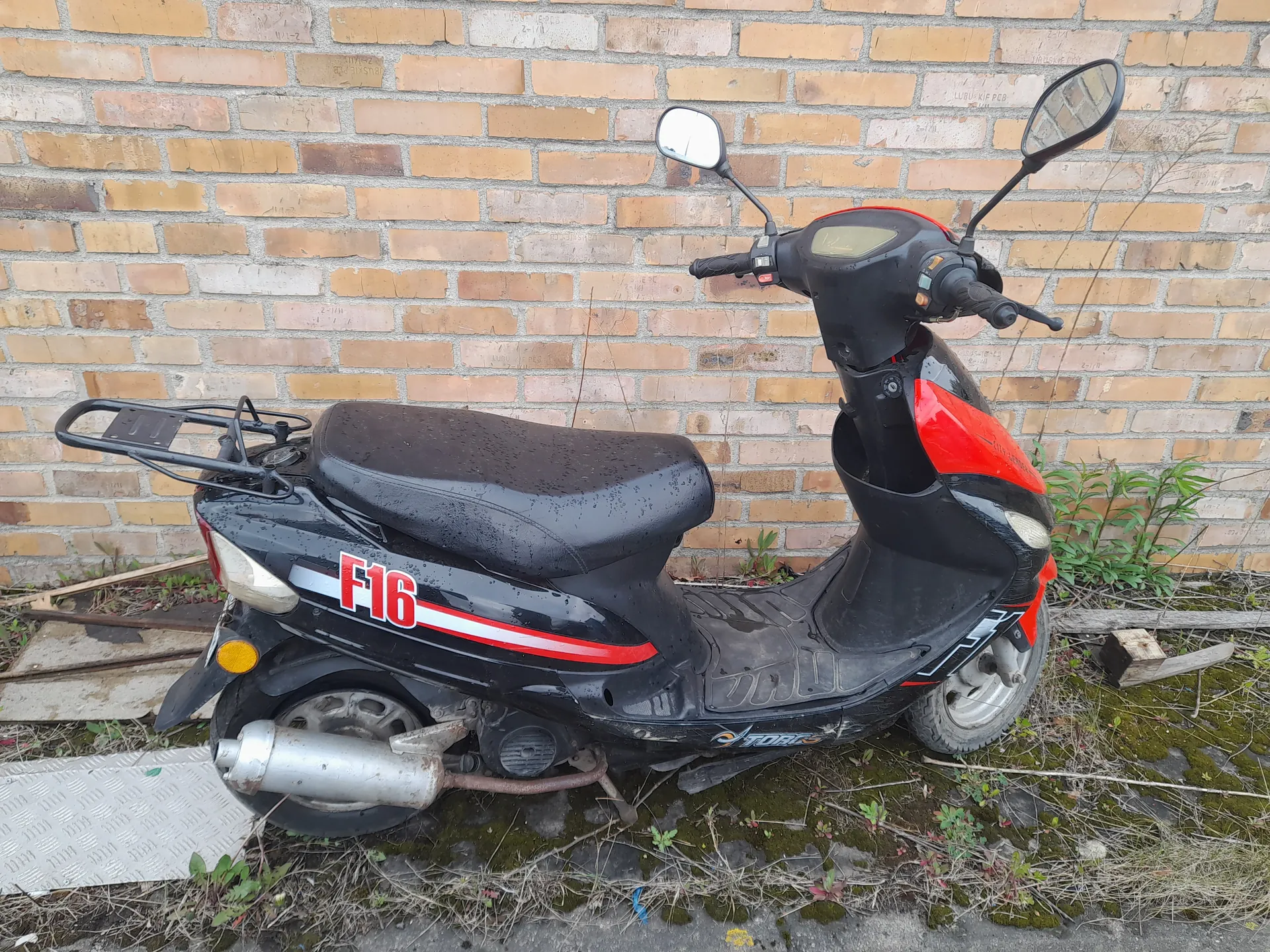 Czarny skuter z czerwonymi akcentami zaparkowany przed ceglaną ścianą. Na boku skutera widoczny czerwony napis F16. Skuter posiada czarne siedzenie, dwa lusterka boczne, tylny bagażnik oraz wydech. Pod skutrem rośnie trawa i mchy, a na ziemi leżą deski i metalowa płyta. Ściana z cegieł w tle jest poziomo ułożona, a horyzont jest prawidłowo zorientowany.