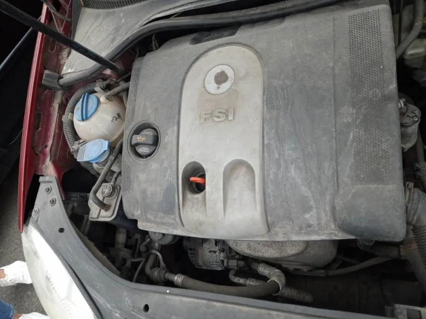 Widok otwartej maski samochodu z silnikiem. Na pokrywie silnika widoczny napis "FSI". Obecne są niebieskie korki zbiorników (chłodniczy i hamulcowy), przewody i elementy mechaniczne. Czerwona część nadwozia widoczna po lewej stronie. Silnik i elementy są umieszczone w standardowej orientacji.