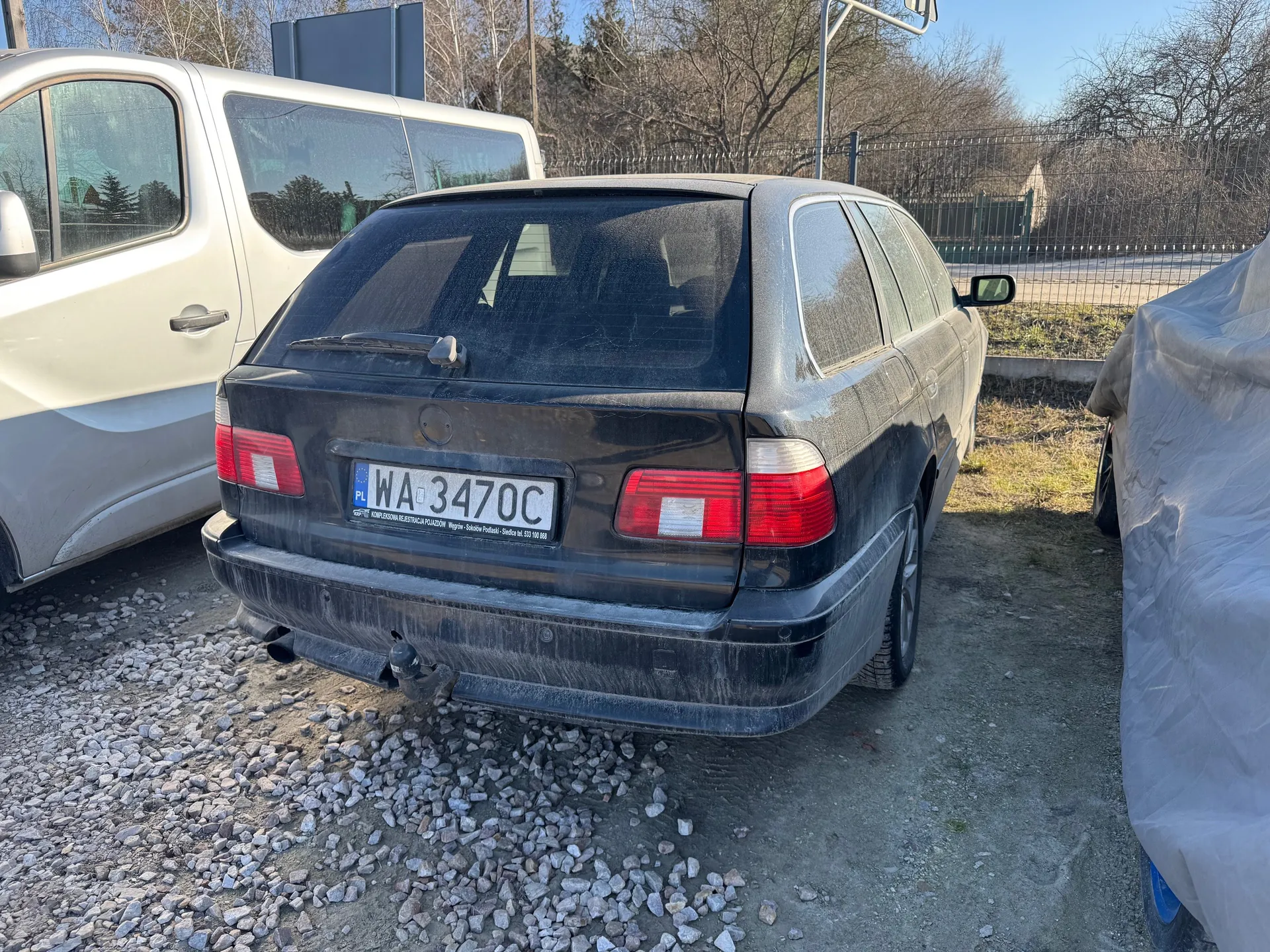 Czarny samochód osobowy typu kombi z tablicą rejestracyjną PL WA 3470C, widok od tyłu. Na tablicy widoczny napis "KOMPLEKSOWA REJESTRACJA POJAZDÓW". Po lewej stronie biały furgon, po prawej zakryty pojazd. Tło: drzewa bez liści, ogrodzenie metalowe, droga i niebo. Podłoga z żwiru i ziemi.