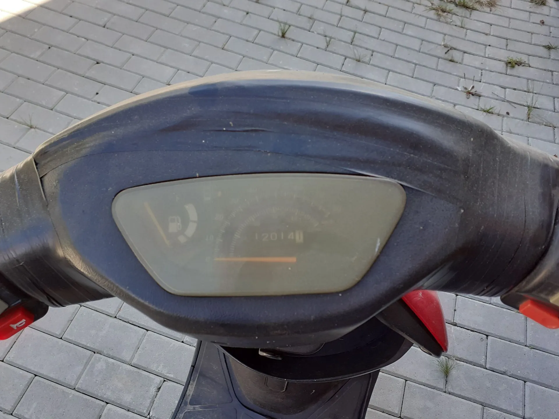 Zbliżenie na deskę rozdzielczą czarnego skutera. Widoczny jest wskaźnik poziomu paliwa oraz licznik kilometrów z wartością 12014. Całość obudowy wokół licznika jest owinięta czarną taśmą izolacyjną. Tło stanowi szara kostka brukowa.