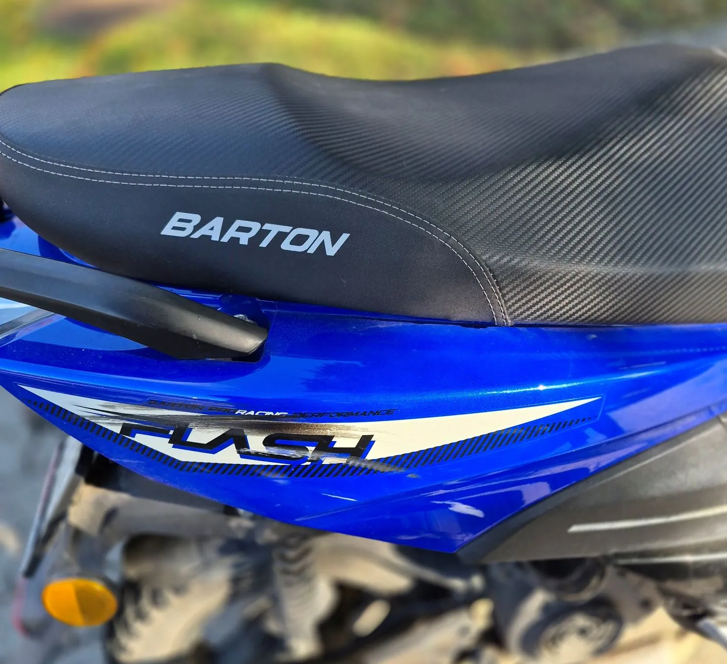 Zbliżenie na tylną część niebieskiego motocykla. Widoczne jest czarne, teksturowane siedzenie z białym napisem „BARTON”. Poniżej, na niebieskiej owiewce, znajduje się grafika z napisem „FLASH” oraz mniejszym tekstem „RACING PERFORMANCE”. W dolnej części obrazu widoczny jest pomarańczowy kierunkowskaz oraz elementy ramy i zawieszenia motocykla.