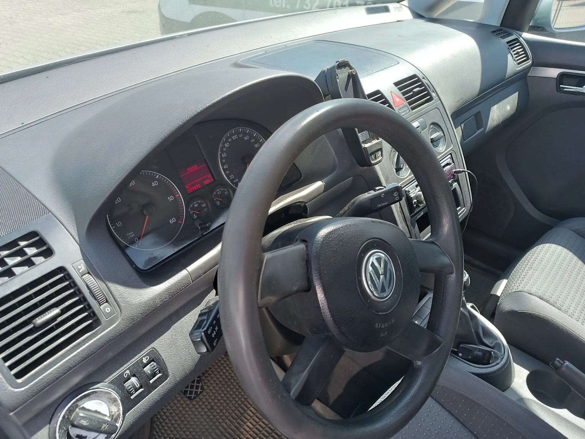 Wnętrze samochodu marki Volkswagen, widok z perspektywy kierowcy. W centrum znajduje się czarna kierownica z logo VW. W tle widoczna deska rozdzielcza z analogowymi zegarami prędkości i obrotomierzem oraz cyfrowym wyświetlaczem przebiegu. Po prawej stronie znajdują się otwory wentylacyjne i panel centralny. Widoczny jest fragment szarego fotela pasażera oraz lewarek zmiany biegów.
