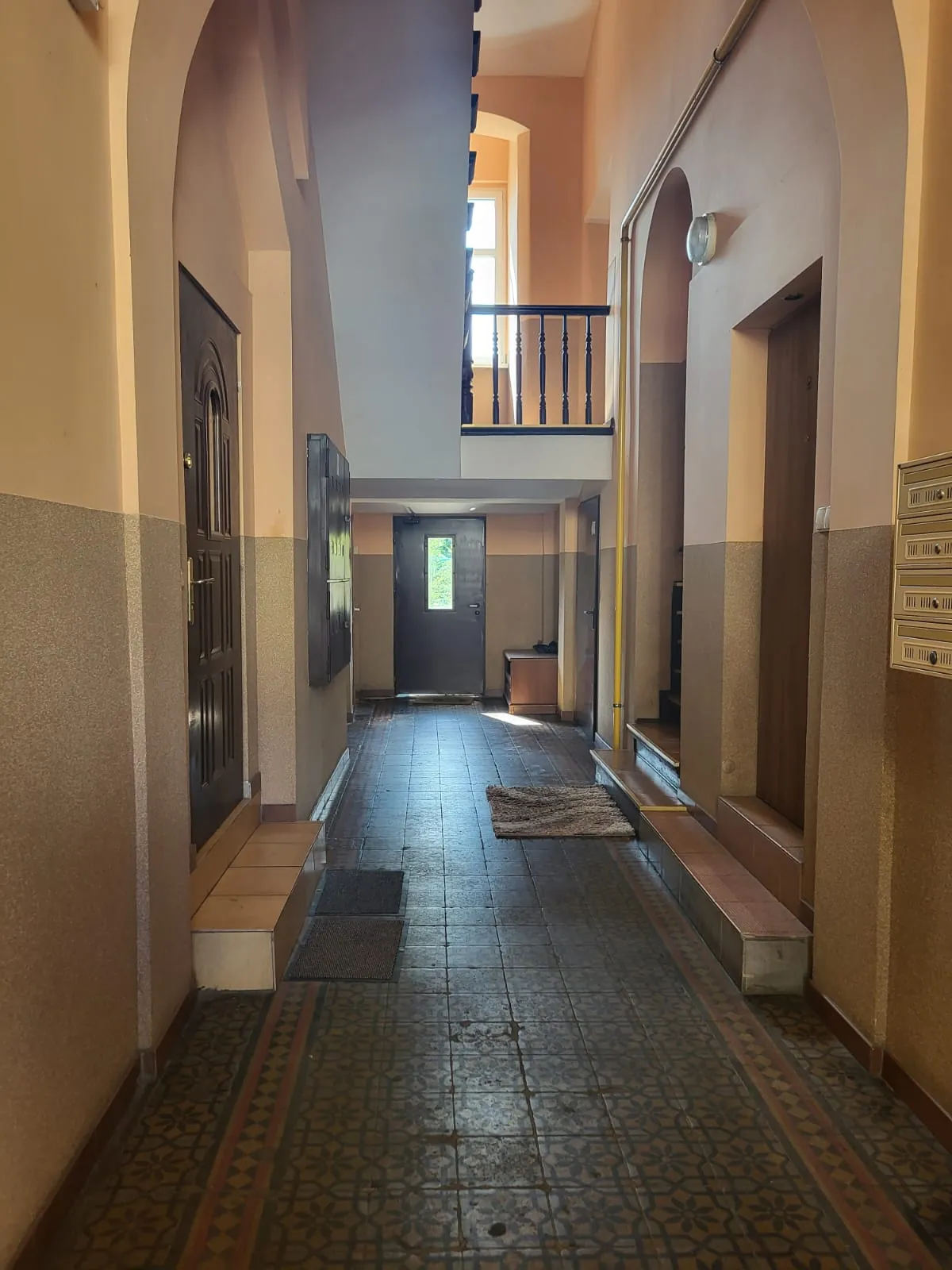 Wnętrze korytarza w budynku o beżowych ścianach. Po lewej stronie znajdują się ciemnobrązowe drzwi i szara szafka elektryczna. Podłoga wyłożona jest kafelkami z ozdobnym wzorem w dolnej części i gładkimi płytkami w głębi. Na końcu korytarza widoczne są kolejne drzwi z przeszkleniem. Po prawej stronie znajduje się przejście do innego pomieszczenia oraz zestaw skrzynek pocztowych zamontowanych na ścianie. Nad korytarzem znajduje się antresola z ciemną balustradą i oknem w tle.