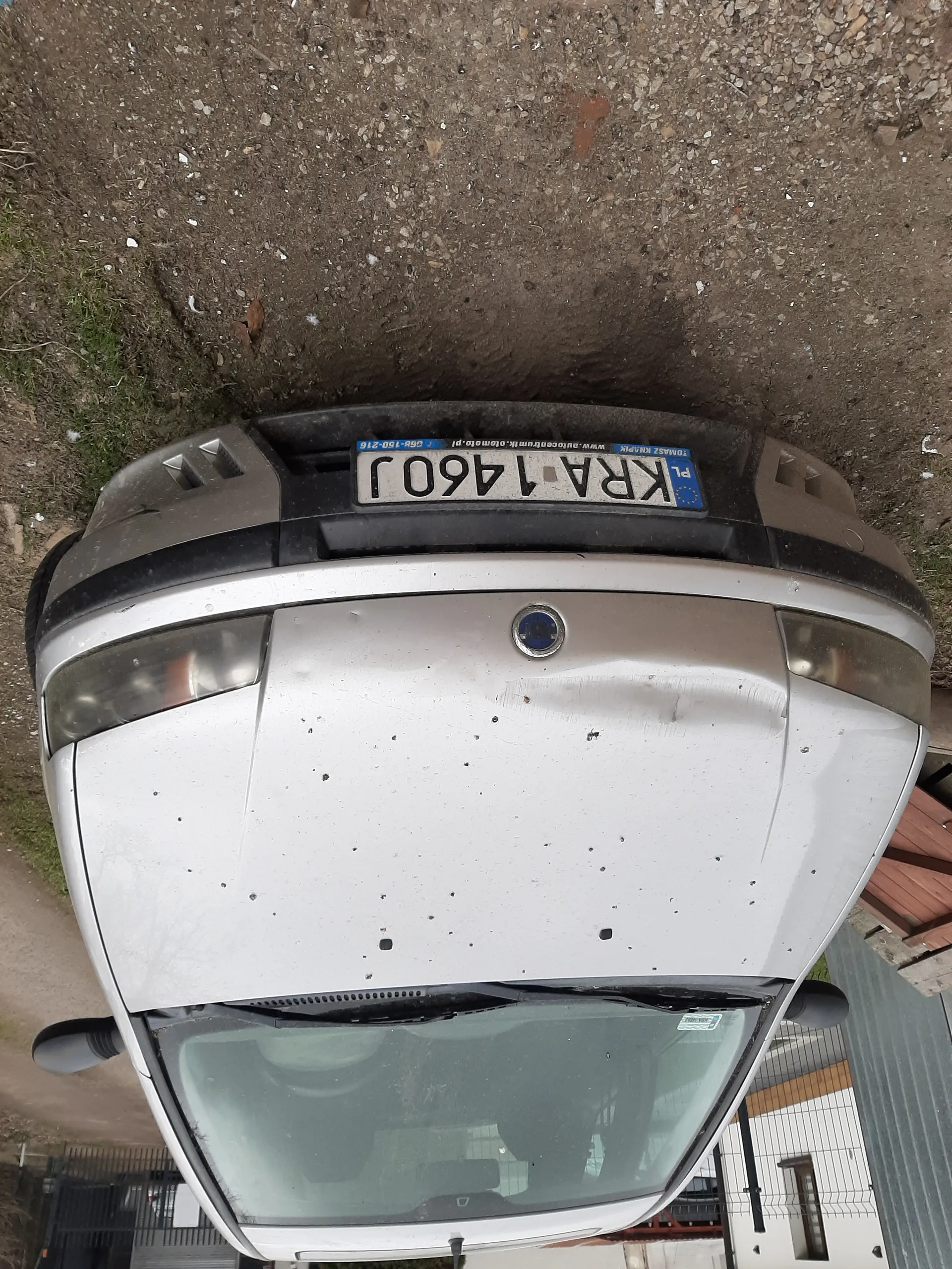 Srebrny samochód widziany od przodu z tablicą rejestracyjną KRA 1460J. Na masce widoczne zadrapania i wgniecenia. Samochód zaparkowany na żwirowym podłożu z trawą w tle. W tle budynek z białą i żółtą elewacją oraz metalowe ogrodzenie.
