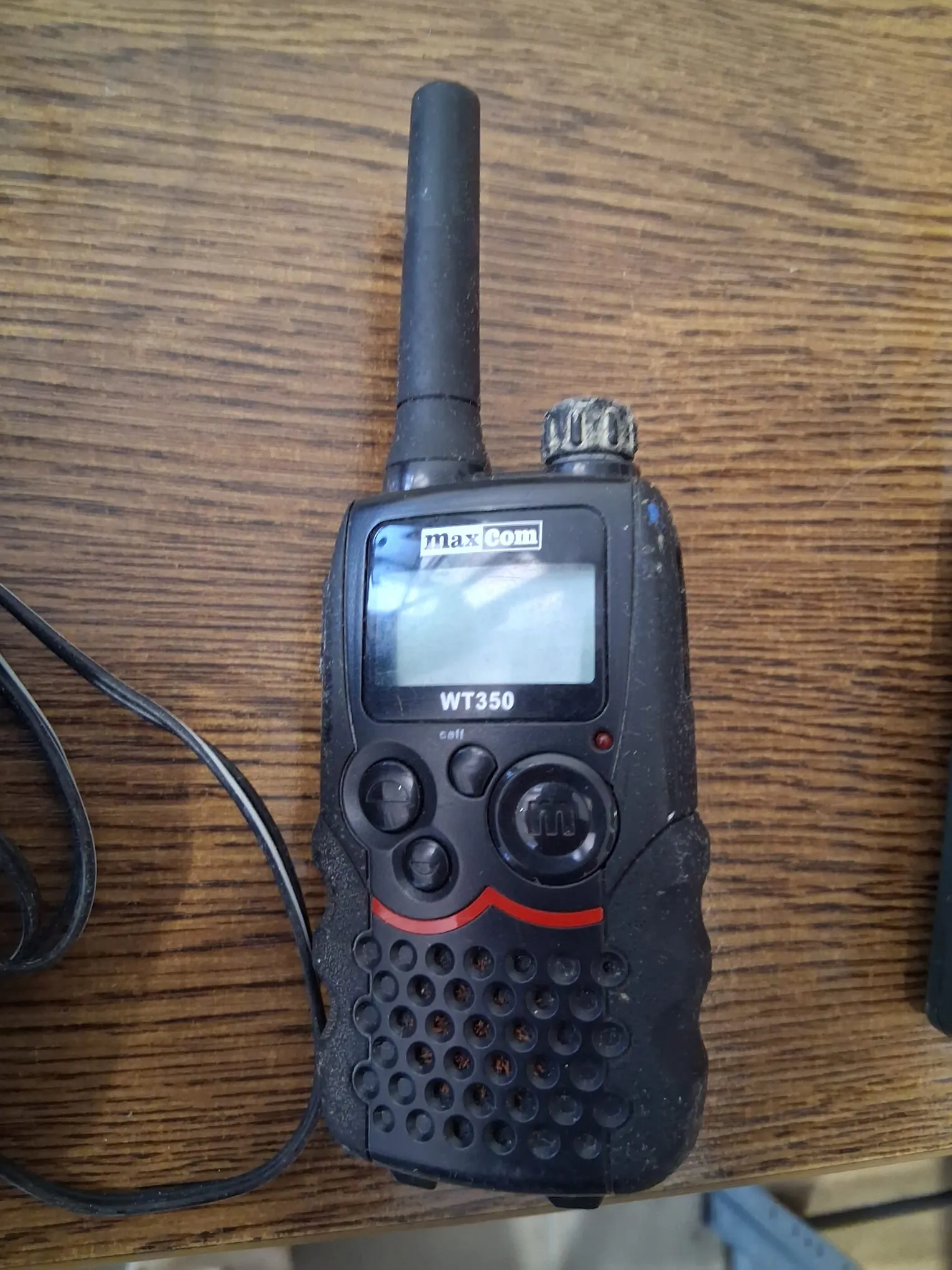 Czarna radiostacja marki MaxCom, model WT350, leżąca na drewnianym blacie. Urządzenie posiada długą antenę na górze, mały ekran LCD, kilka przycisków sterujących oraz perforowaną obudowę w dolnej części. Po lewej stronie widoczny jest czarny kabel.