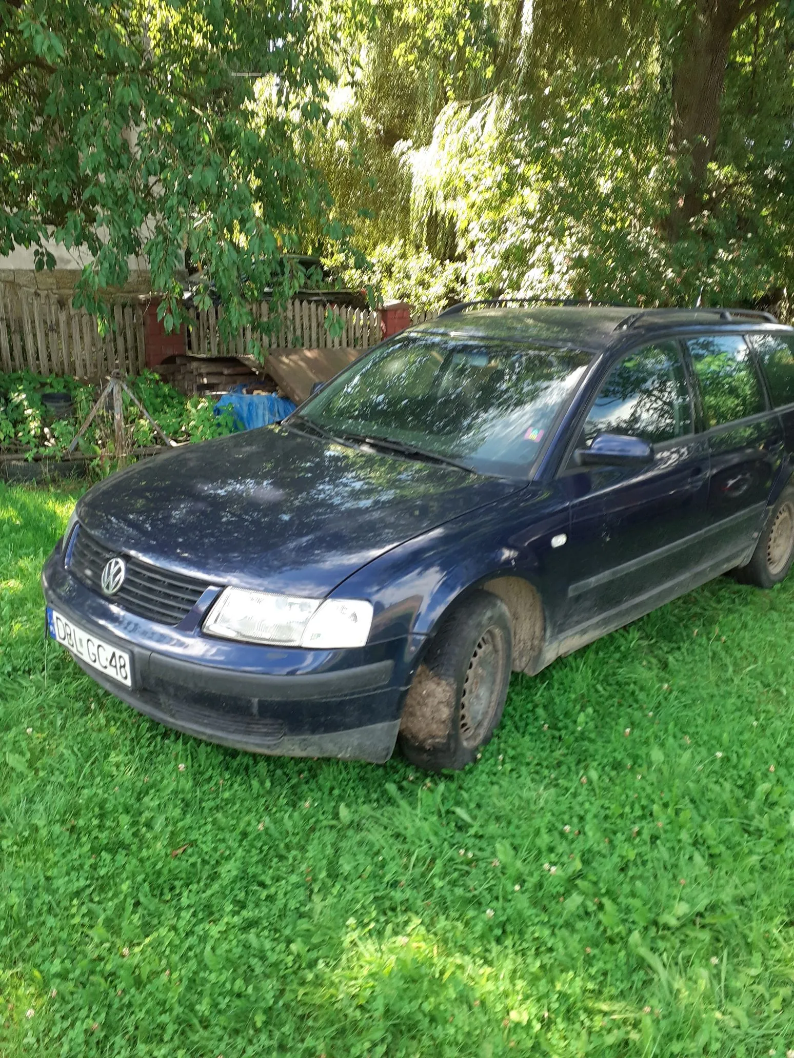 Ciemnoniebieski samochód marki Volkswagen Passat w nadwoziu kombi, zaparkowany na trawie. Pojazd jest brudny, a przednie prawe koło jest częściowo zagłębione w ziemi. Widoczna jest tablica rejestracyjna o numerze CBL GC48. W tle znajduje się drewniany płot oraz gęste, zielone drzewa i krzewy.