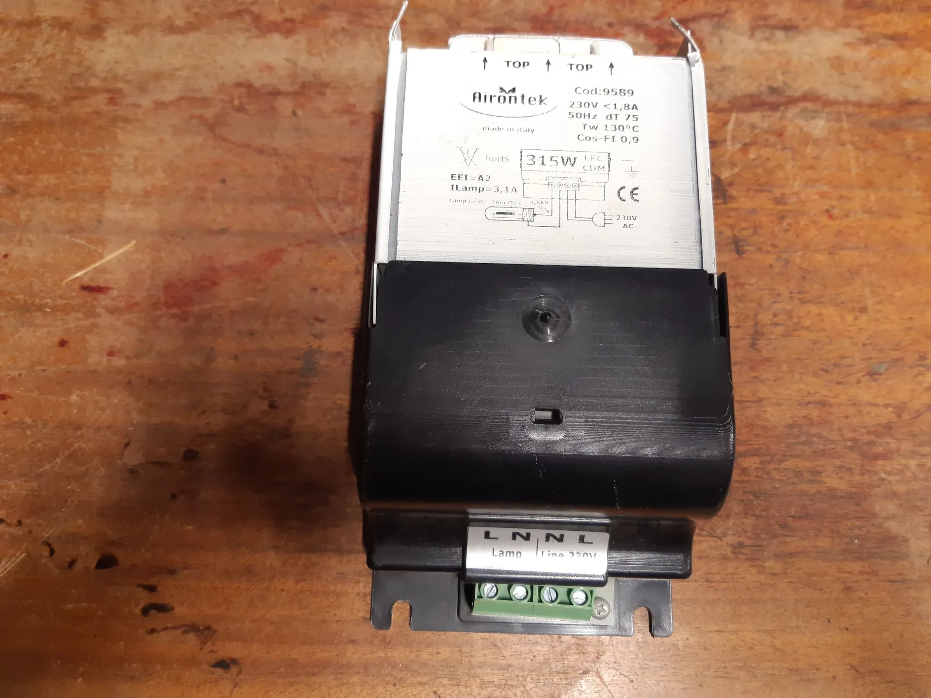 Biały i czarny zasilacz elektroniczny do lampy, leżący na drewnianej powierzchni. Na górnej białej części znajduje się napis 'Airontek' oraz dane techniczne: 'Cod: 9589', '230V < 1,8A', '50Hz', 'Tw 130°C', 'Cos-Fi 0,9', '315W', 'EEI = A2', 'I Lamp = 3,1A'. Widoczny jest również znak CE oraz schemat elektryczny. Na dole urządzenia znajduje się zielona kostka przyłączeniowa z oznaczeniami 'L N N L' oraz 'Lamp Line 230V'.