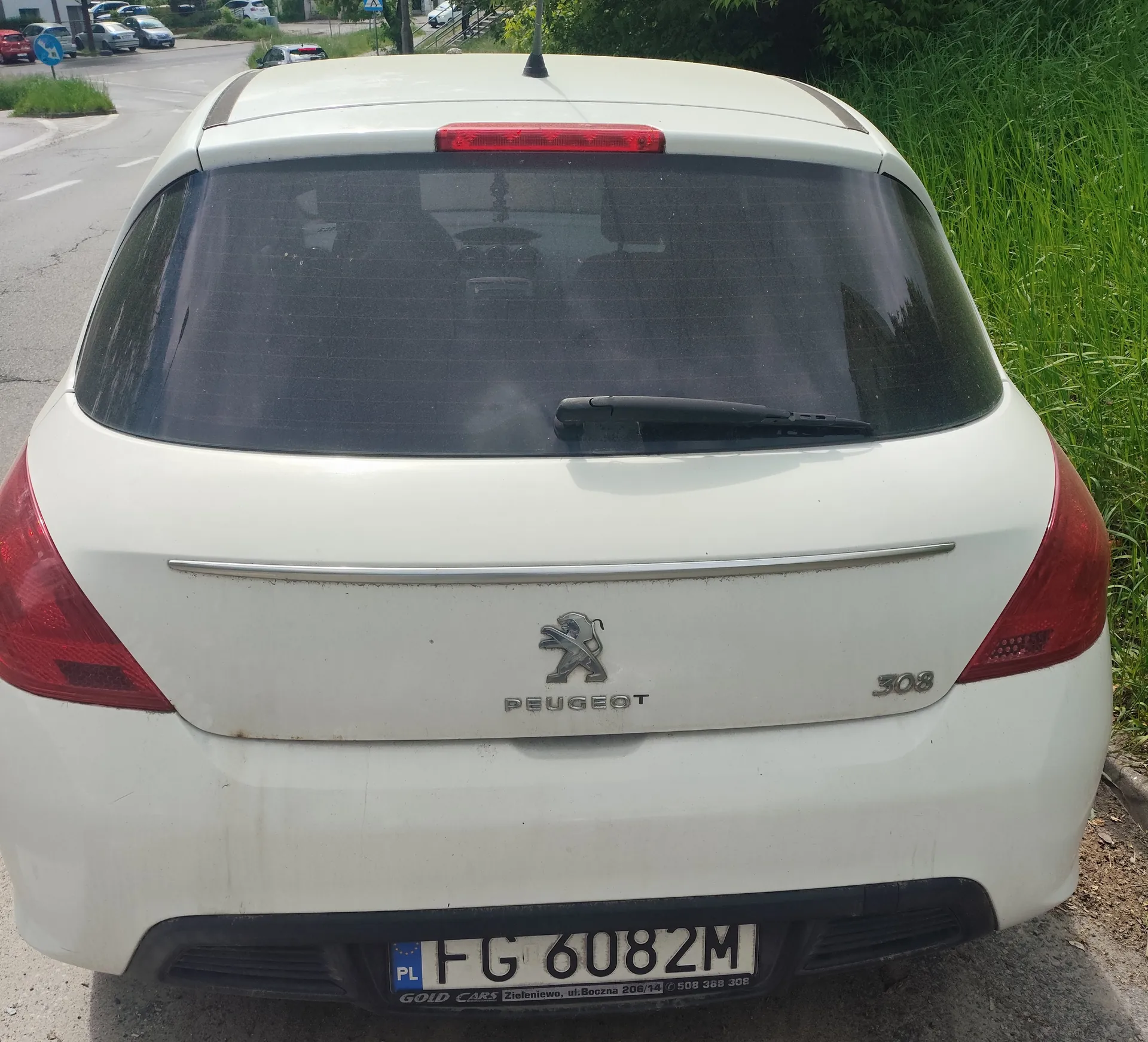 Tył białego samochodu Peugeot 308 zaparkowanego przy drodze. Widoczne są czerwone światła tylnego, szyba z wycieraczką, logo Peugeot oraz oznaczenie modelu 308. Tablica rejestracyjna z polskim kodem PL i numerem FG 6082M. W tle droga z innymi pojazdami, zielona trawa i znak drogowy.
