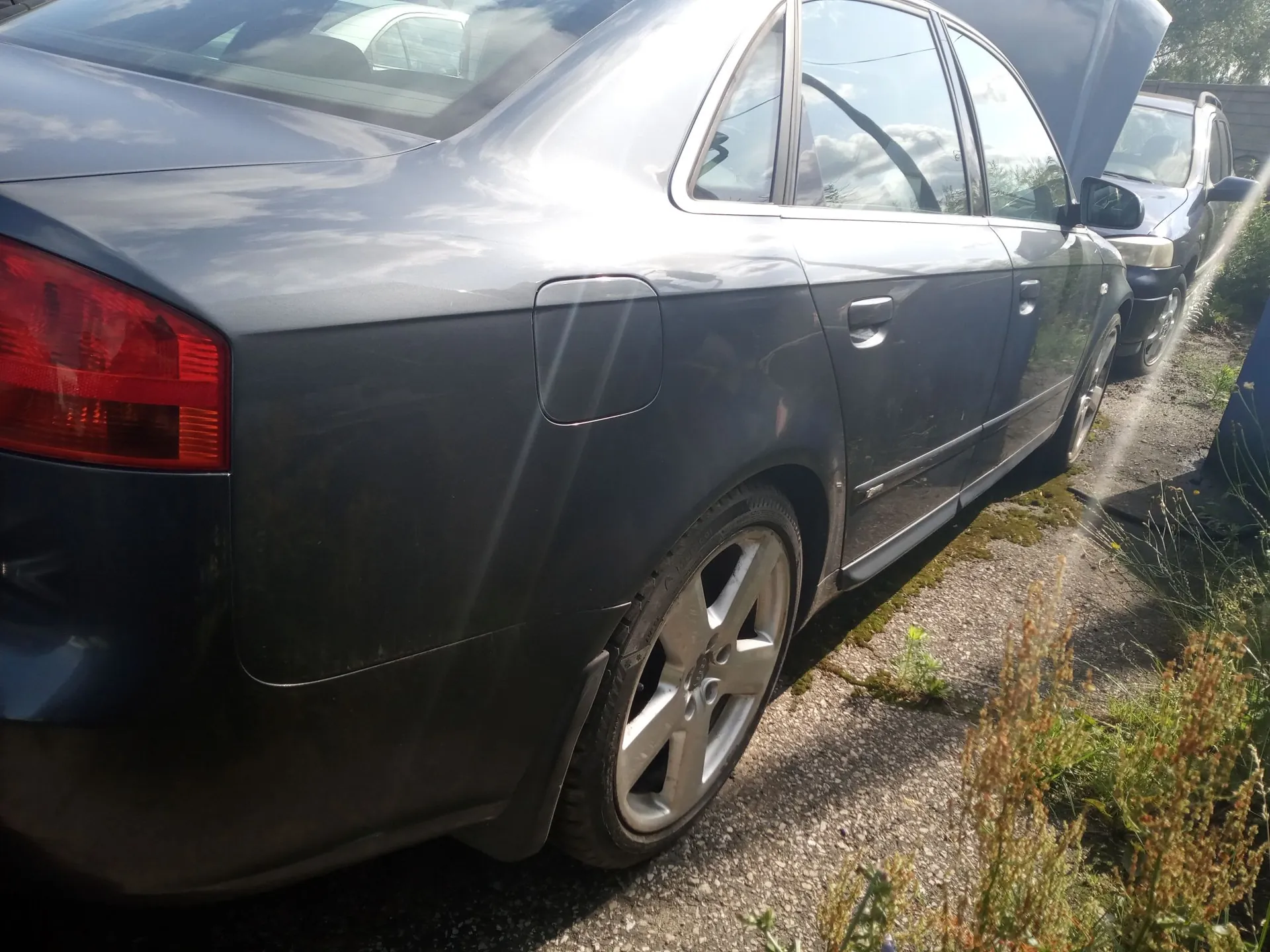 Zdjęcie przedstawiające tylną i boczną część srebrno-szarego samochodu osobowego zaparkowanego na żwirowej drodze. Widoczny jest lewy tylny klosz lampy, klapka wlewu paliwa oraz jedno z kół z pięcioramienną felgą aluminiową. W tle widać fragment innego, ciemniejszego samochodu oraz roślinność w postaci traw i chwastów.