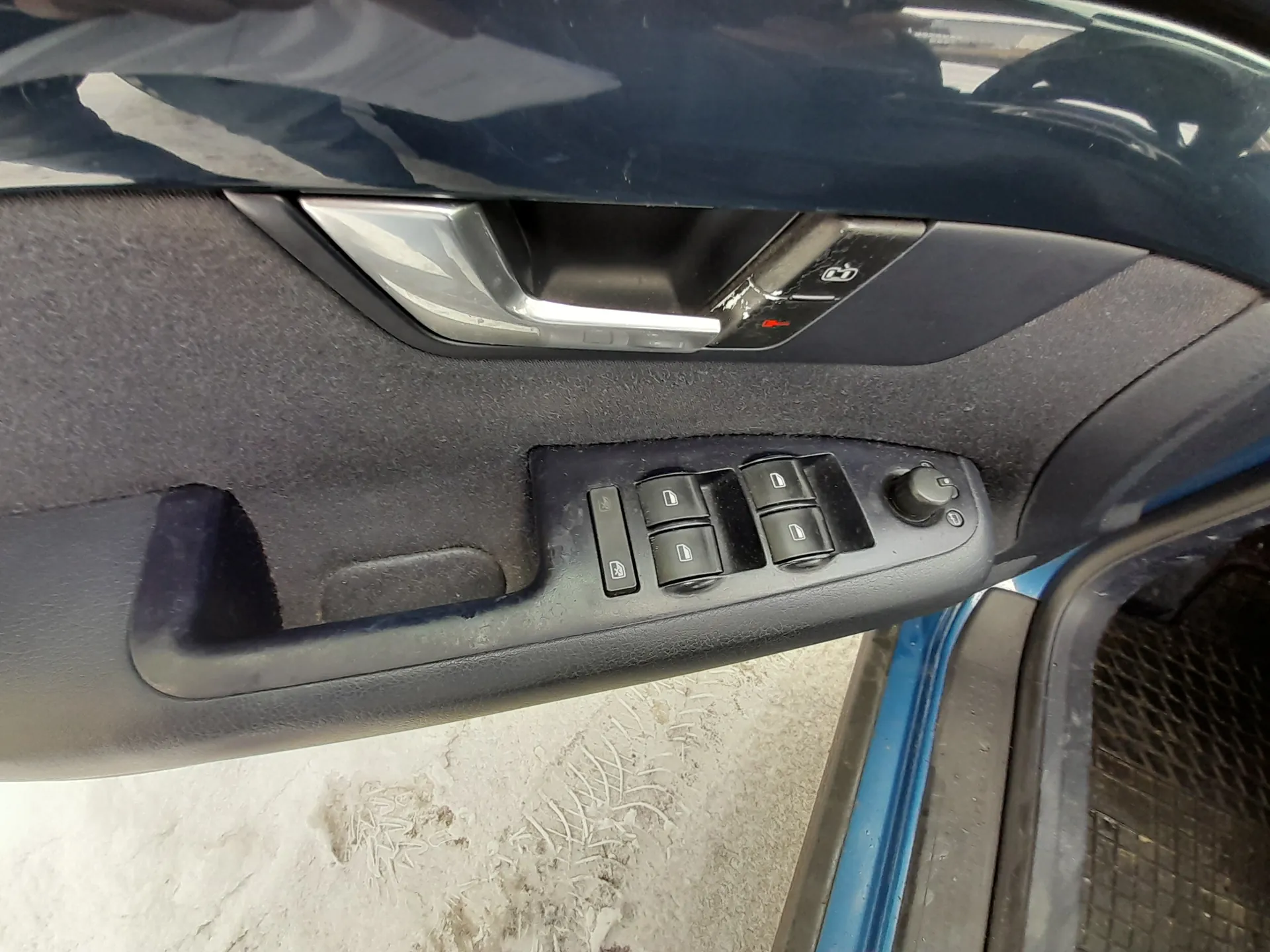 Wnętrze drzwi samochodu z czarnym panelu, srebrną klamką i czarnymi przyciskami sterującymi szybami. Widoczne są cztery przyciski z symbolami okien oraz okrągły regulator. Po prawej stronie fragment niebieskiego nadwozia i siatki. Na zewnątrz śnieg z odciskami.