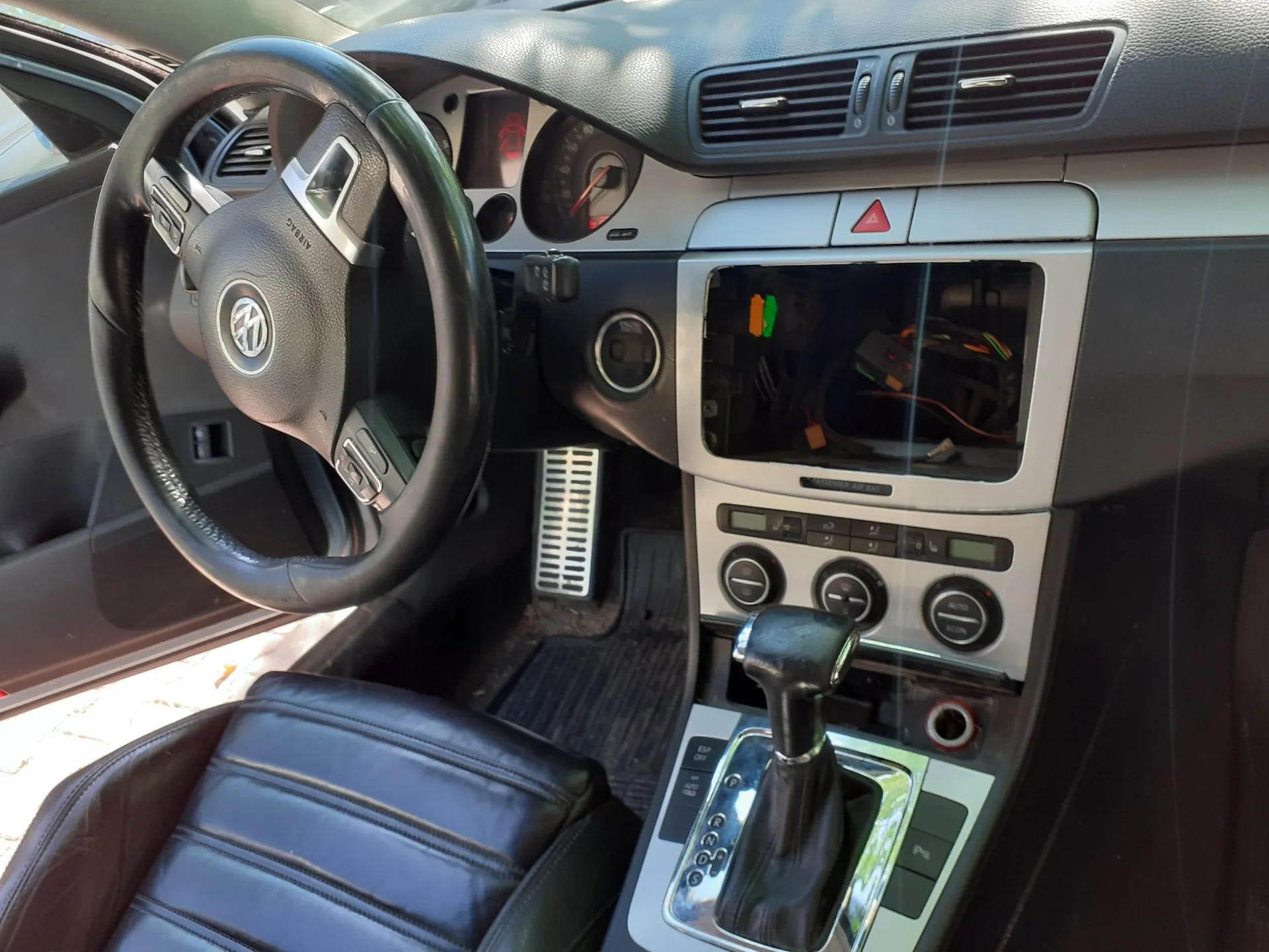 Wnętrze samochodu z perspektywy kierowcy. Widoczna jest czarna skórzana kierownica z logo Volkswagena, deska rozdzielcza z licznikiem oraz konsola środkowa. W miejscu radia znajduje się pusta wnęka z widocznymi przewodami elektrycznymi. Poniżej znajdują się pokrętła klimatyzacji oraz dźwignia zmiany biegów automatycznej. W tle widoczny jest aluminiowy pedał gazu i hamulca oraz czarny skórzany fotel kierowcy.