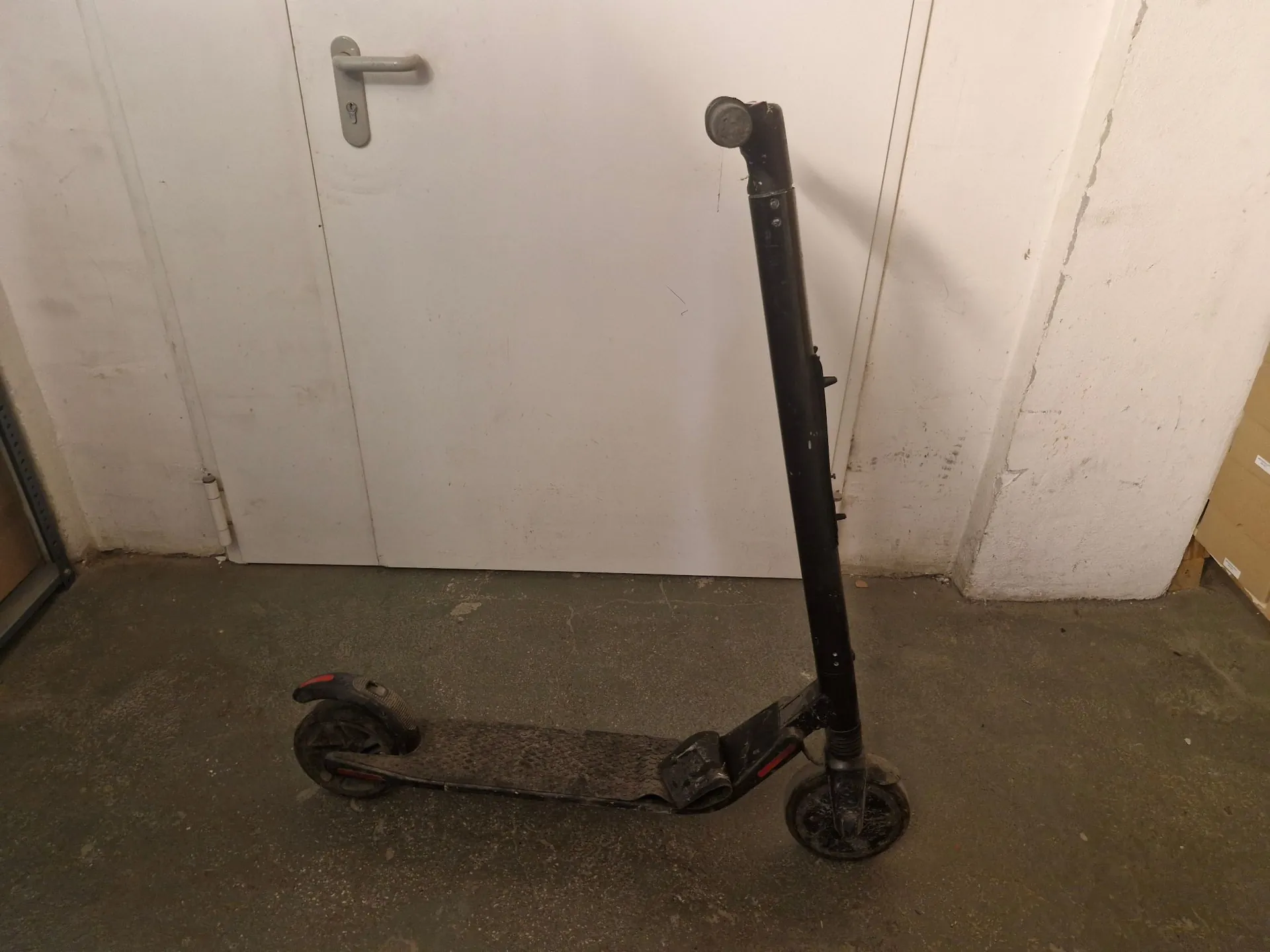Czarny scooter elektryczny stojący na szarym, betonowym podłożu przed białymi drzwiami.