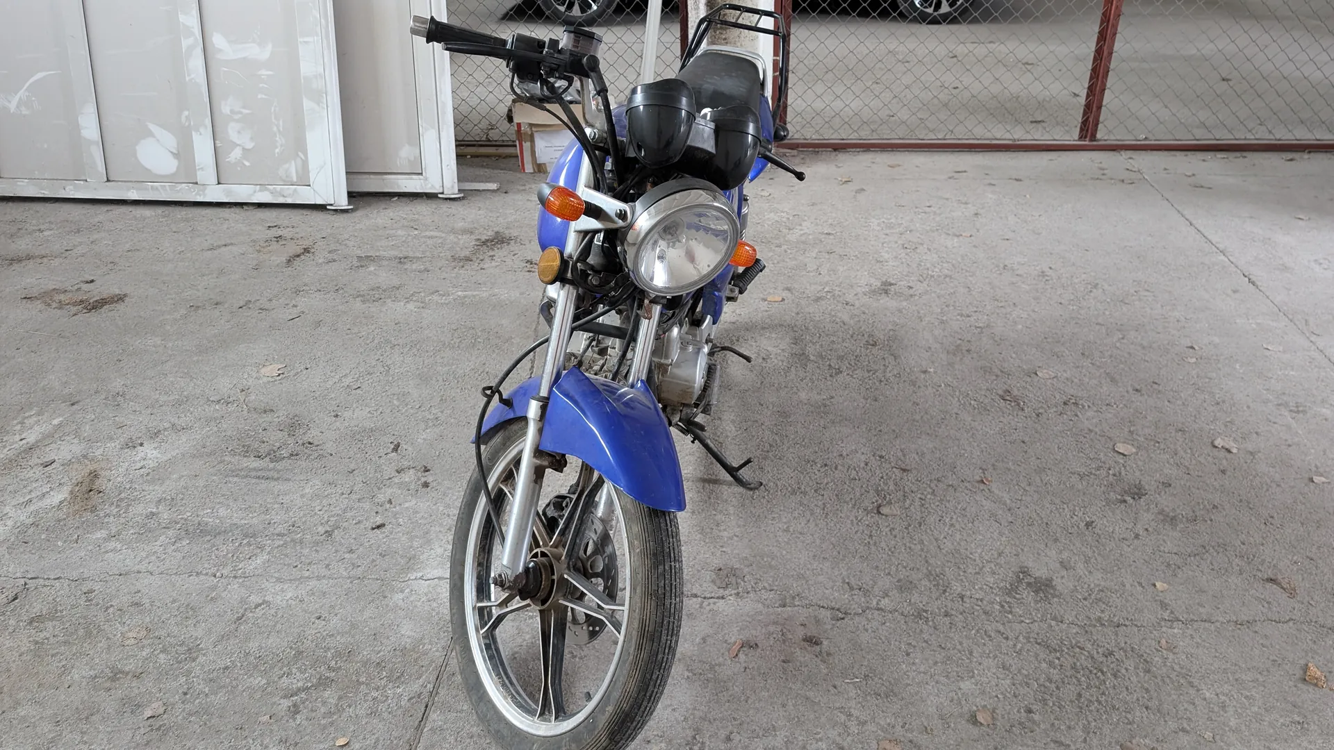 Niebieski motocykl z przodem skierowanym do przodu, z dużym reflektorem, pomarańczowymi kierunkowskazami i czarnym siedzeniem. Motocykl stoi na betonowej powierzchni z widocznymi plamami i liśćmi. W tle siatka ogrodzeniowa, białe metalowe panele z odłupaną farbą oraz fragment koła samochodu za ogrodzeniem.