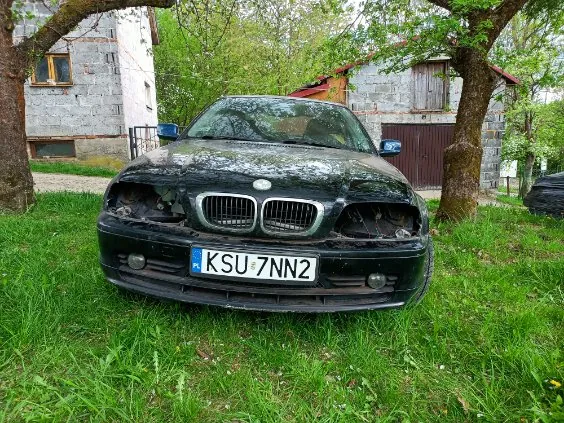 Zniszczony czarny samochód marki BMW z tablicą rejestracyjną K SU 7NN2, stojący na trawie. W tle budynki z kamienia i drzewa. Przednia część pojazdu uszkodzona, brak reflektorów.
