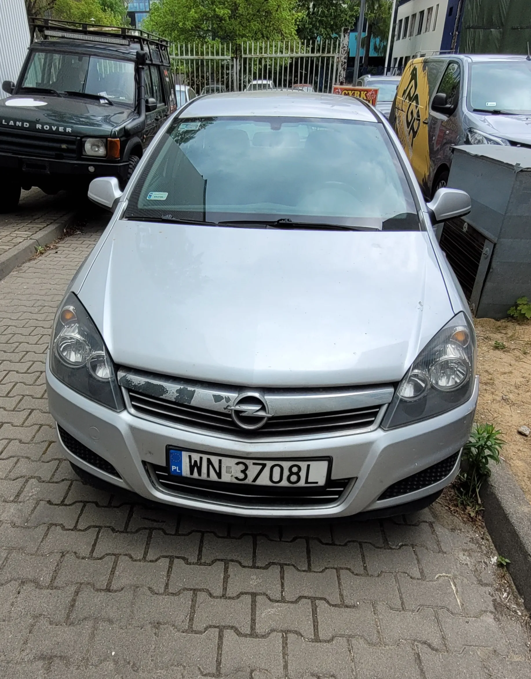Przedni widok srebrnego samochodu marki Opel z tablicą rejestracyjną PL WN 3708L. Po lewej stronie czarny samochód Land Rover, po prawej żółty van z czarnym wzorem. Tło: metalowe ogrodzenie, drzewa, budynki i inne zaparkowane pojazdy. Samochody stoją na brukowanej powierzchni.