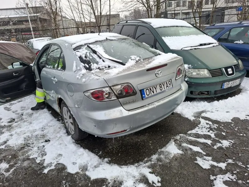 Stojące na śnieżnym parkingu samochody. W centrum srebrny samochód marki Mazda z tablicą rejestracyjną ONY 81A91, na dachu i szybie tylnej śnieg, otwarte drzwi kierowcy. Obok niego zielony samochód marki SEAT z tablicą 08 6891. W tle budynki, drzewa bez liści i inne zaparkowane pojazdy. Na ziemi śnieg i odsłonięta nawierzchnia parkingu.