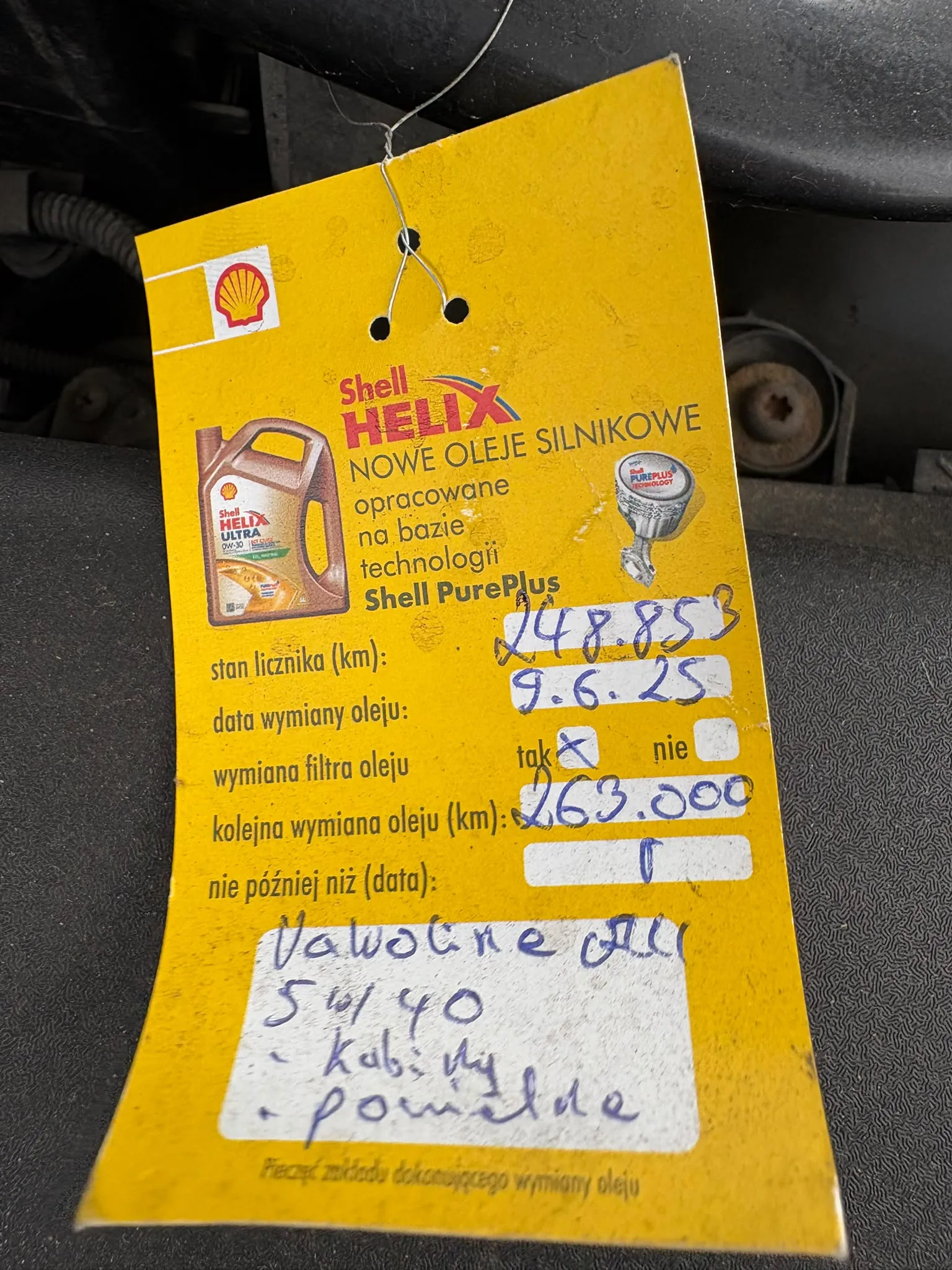 Żółta etykieta serwisowa Shell Helix przyklejona do elementu samochodu. Tekst na etykiecie zawiera napisy: 'Shell HELIX', 'NOWE OLEJE SILNIKOWE opracowane na bazie technologii Shell PurePlus'. Widoczne są pola do wypełnienia: 'stan licznika (km):' z wpisaną wartością '248 853', 'data wymiany oleju:' z wpisaną datą '9.6.25', 'wymiana filtra oleju:' z zaznaczonym polem 'tak', 'kolejna wymiana oleja (km):' z wpisaną wartością '263 000' oraz 'nie później niż (data):'. Po prawej stronie znajdują się odręczne notatki w języku obcym. Etykieta jest przymocowana za pomocą cienkiego drutu przechodzącego przez otwór.