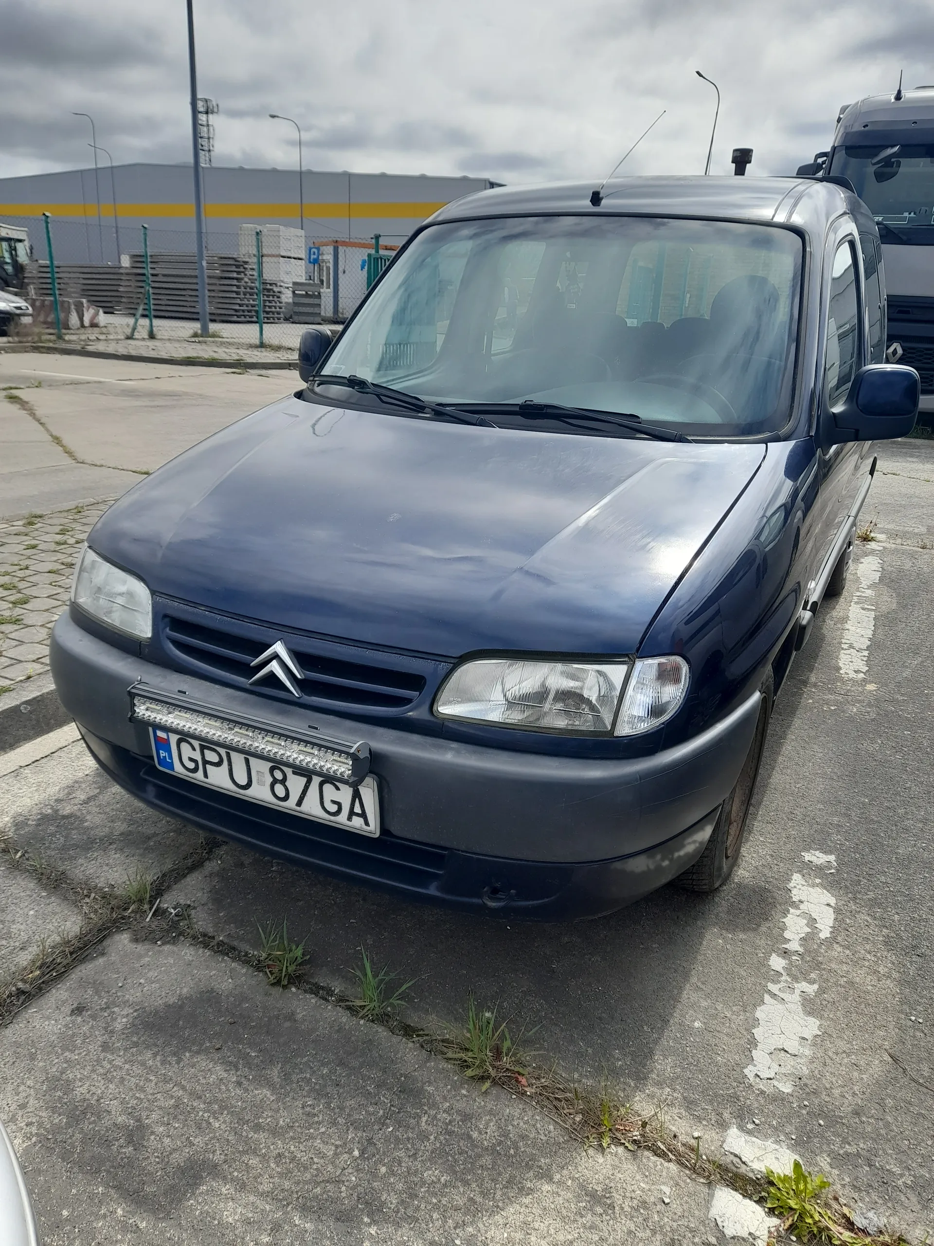 Ciemnoniebieski samochód marki Citroën zaparkowany na asfaltowej powierzchni. Widoczna tablica rejestracyjna z numerem GPU 87GA. W tle drugi pojazd, budynek z żółtą paską, latarnie uliczne oraz chmurne niebo. Na drodze białe linie i niewielkie rośliny przy krawężniku.