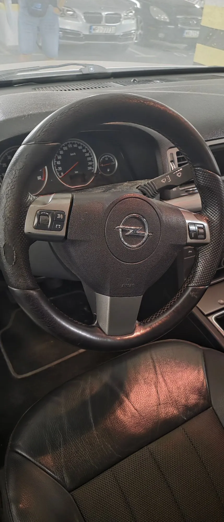 Widok z wnętrza samochodu na kierownicę marki Opel z logo na środku. Kierownica jest obita czarną skórą, która wykazuje oznaki zużycia i przetarcia po bokach. W tle widoczny jest panel wskaźników z prędkościomierzem oraz przednia szyba, przez którą widać inne zaparkowane samochody.
